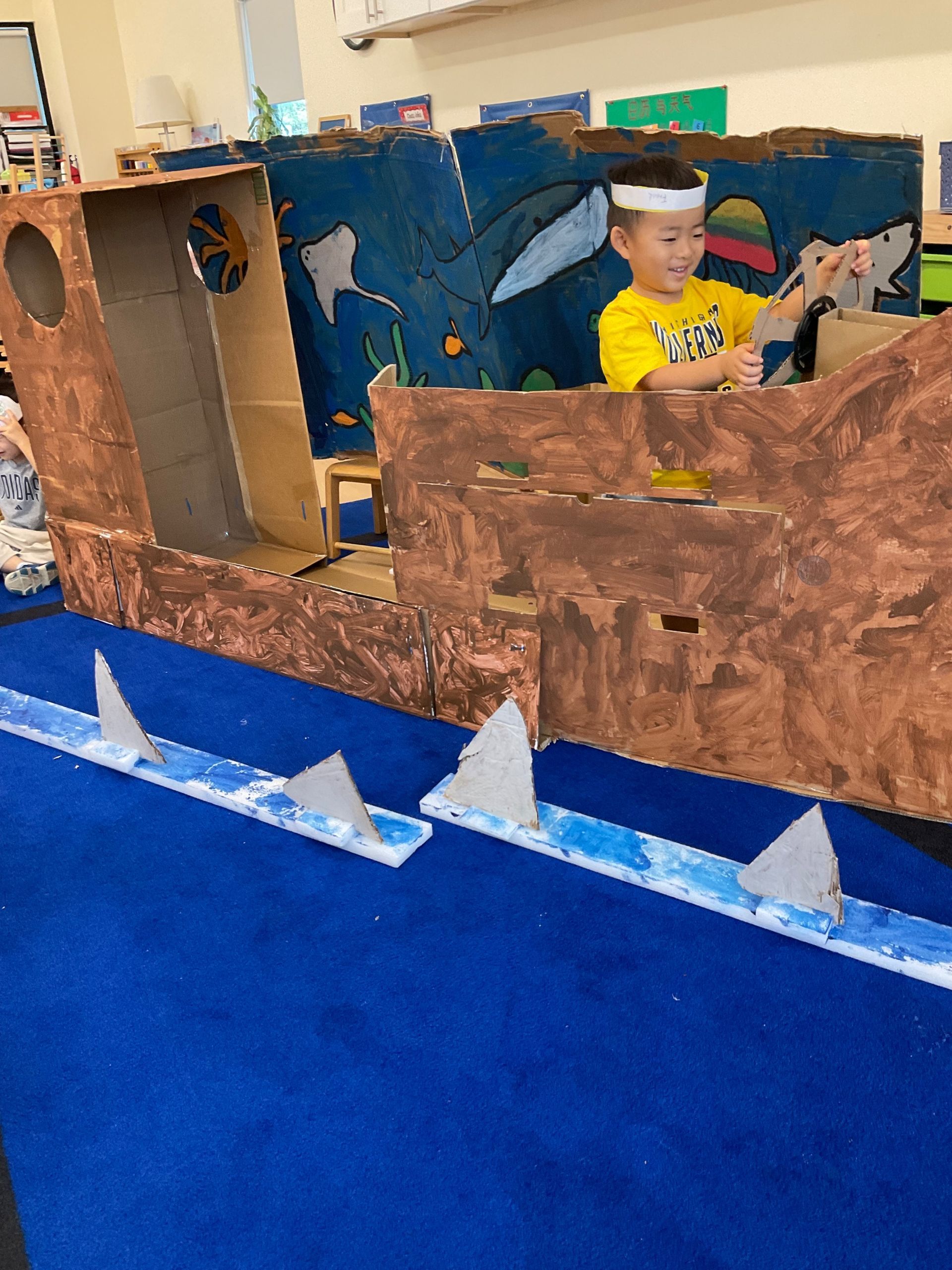 Child in a cardboard pirate ship craft, painted brown and blue, with white waves in a classroom.