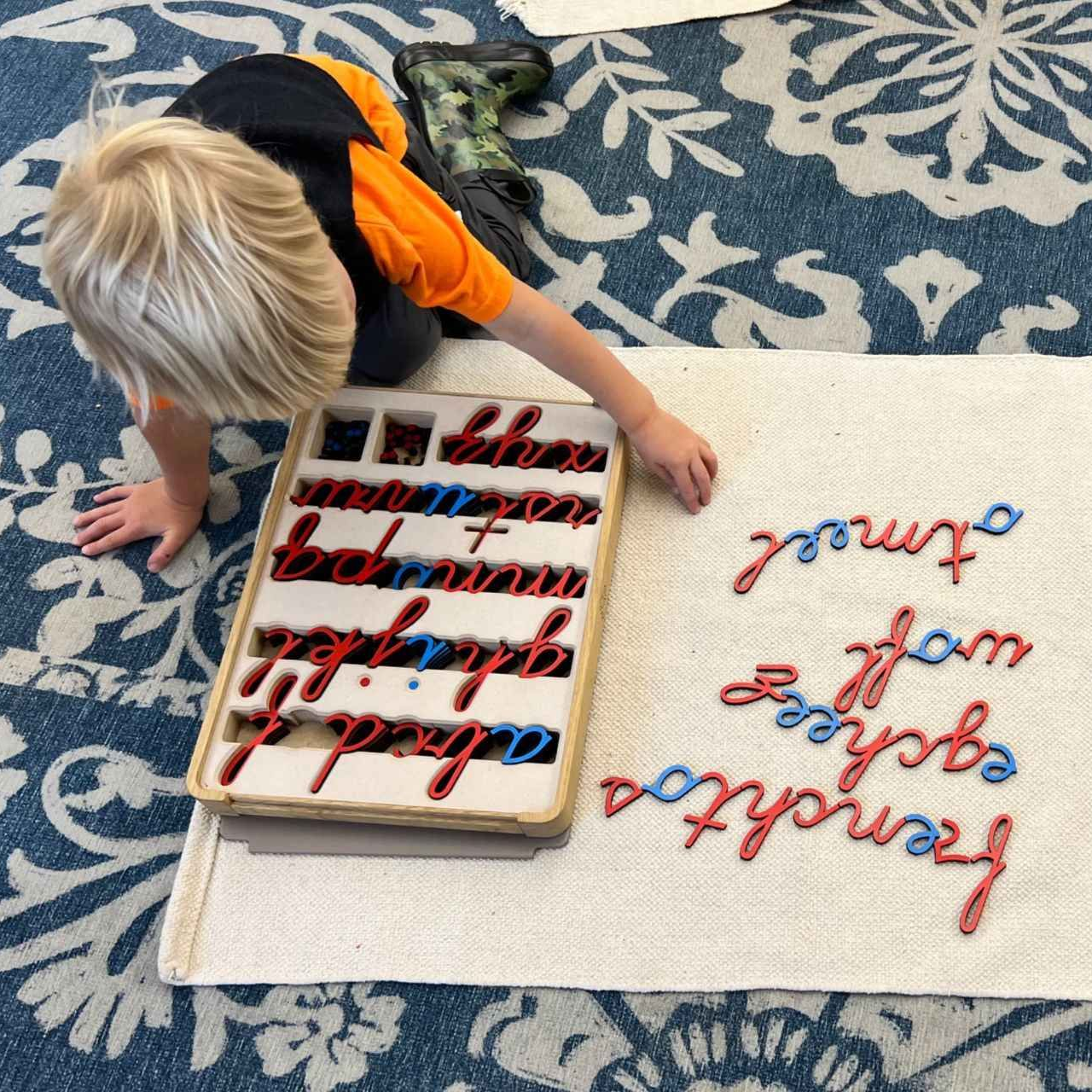 Child using a tray of red and blue letters to spell words on a rug.