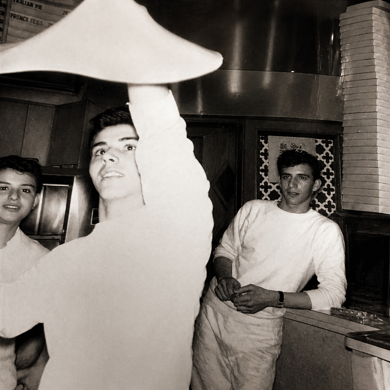 Three kitchen workers in white uniforms preparing food, one reaching up with a tray