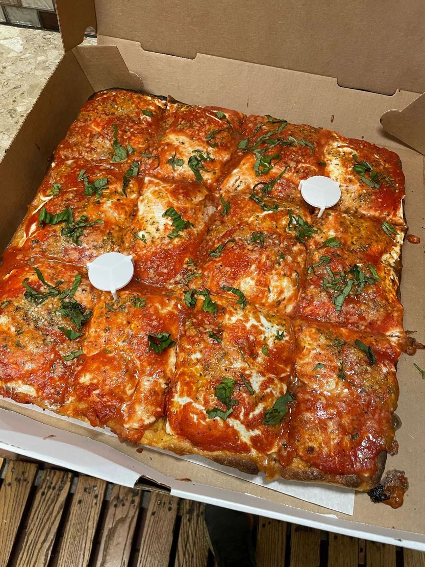 Square pepperoni pizza in a cardboard box, topped with green herbs and two white sauce cups.