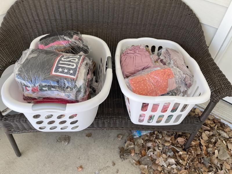 Two white laundry baskets filled with clothes wrapped in clear plastic on a wicker bench.