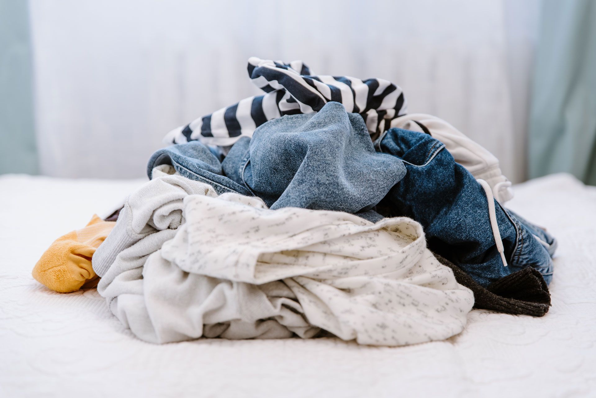 Pile of assorted laundry on a white bedspread; includes jeans, striped shirt, and other clothing items.