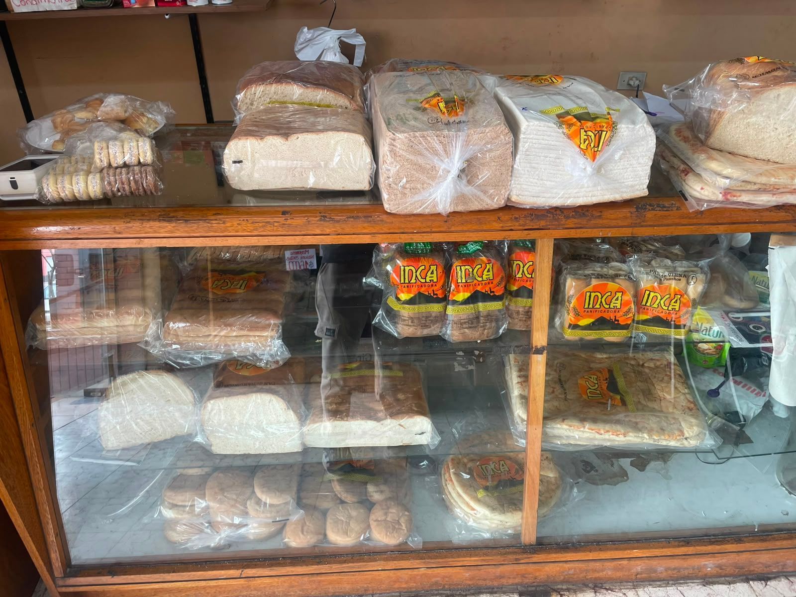 Estantería de panadería con pan y pasteles envueltos, expuestos tras un cristal y sobre un mueble de madera.