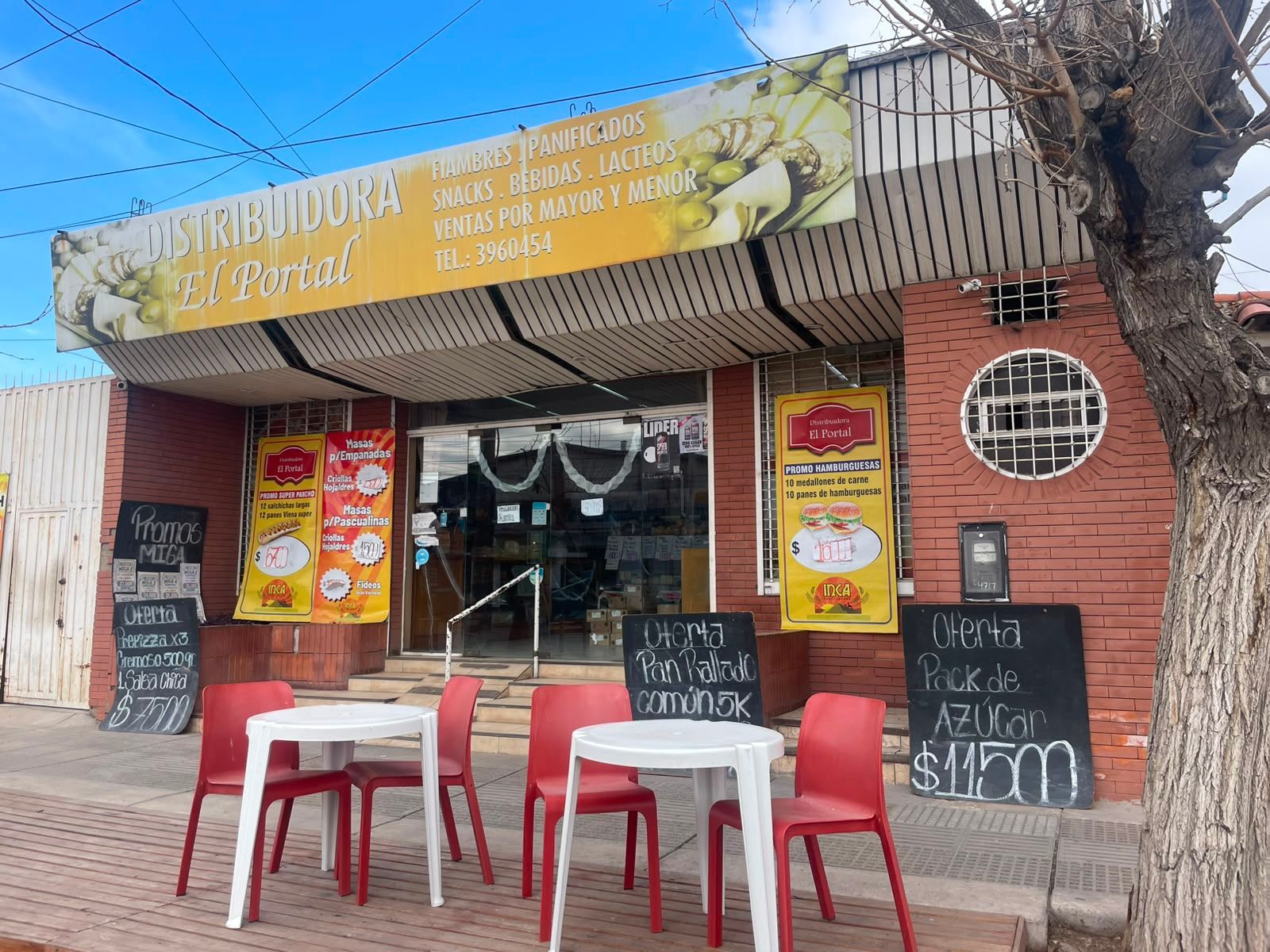 Pequeña cafetería con mesas al aire libre, sillas rojas y carteles con el menú bajo un toldo amarillo.
