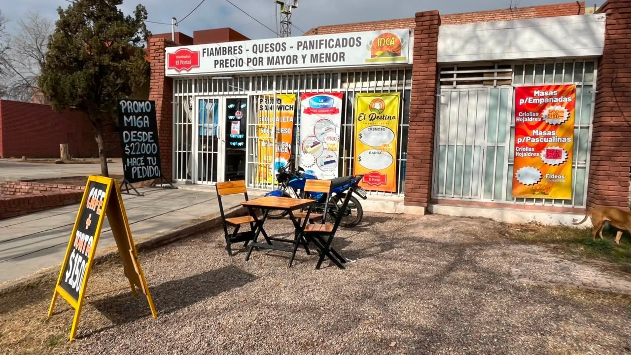 Cafetería al aire libre con toldo naranja, escaparate de cristal y una pequeña mesa con sillas en una zona de grava.