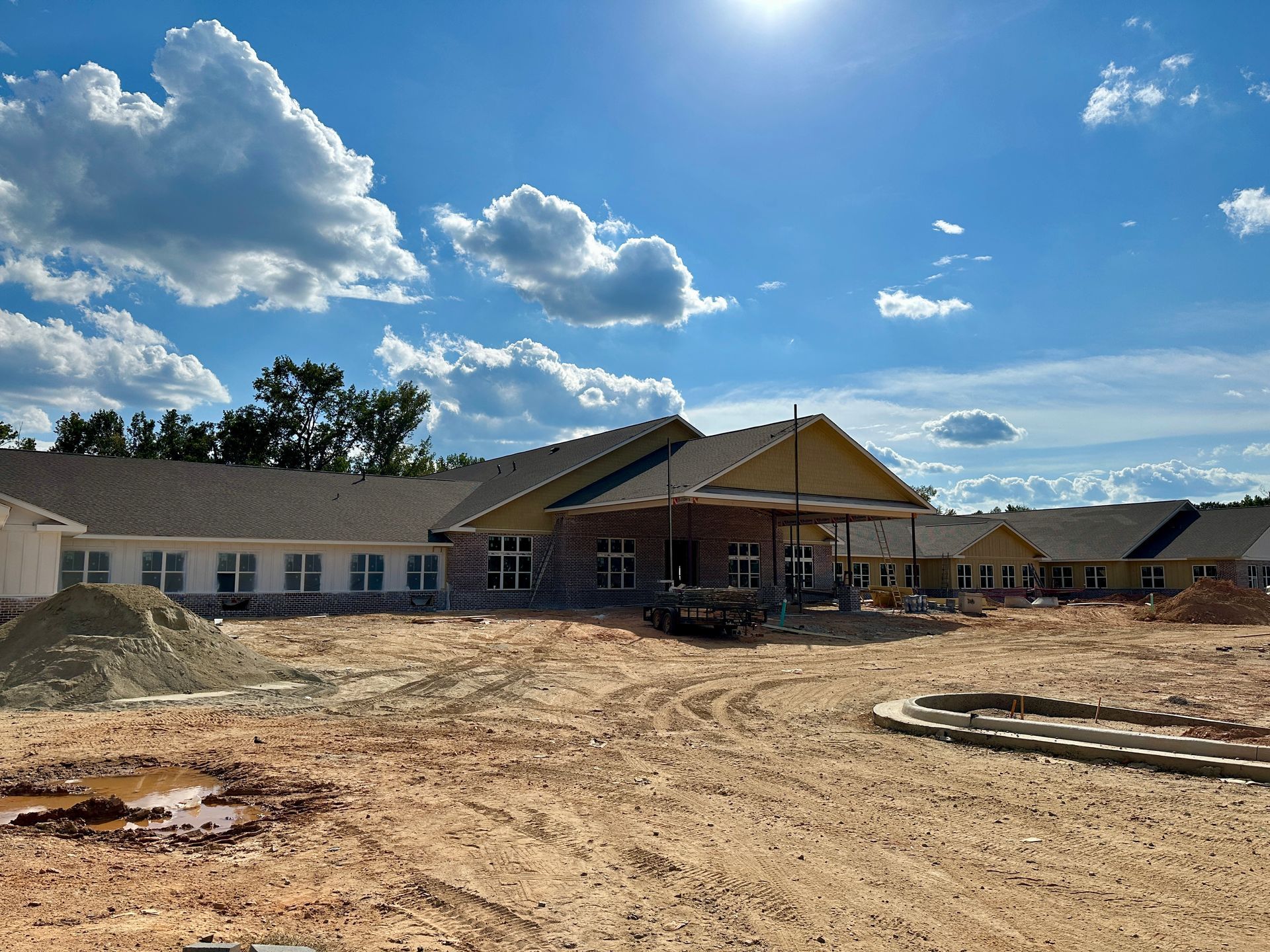 Construction site of a long, one-story building with a covered entrance, against a blue sky with clouds.