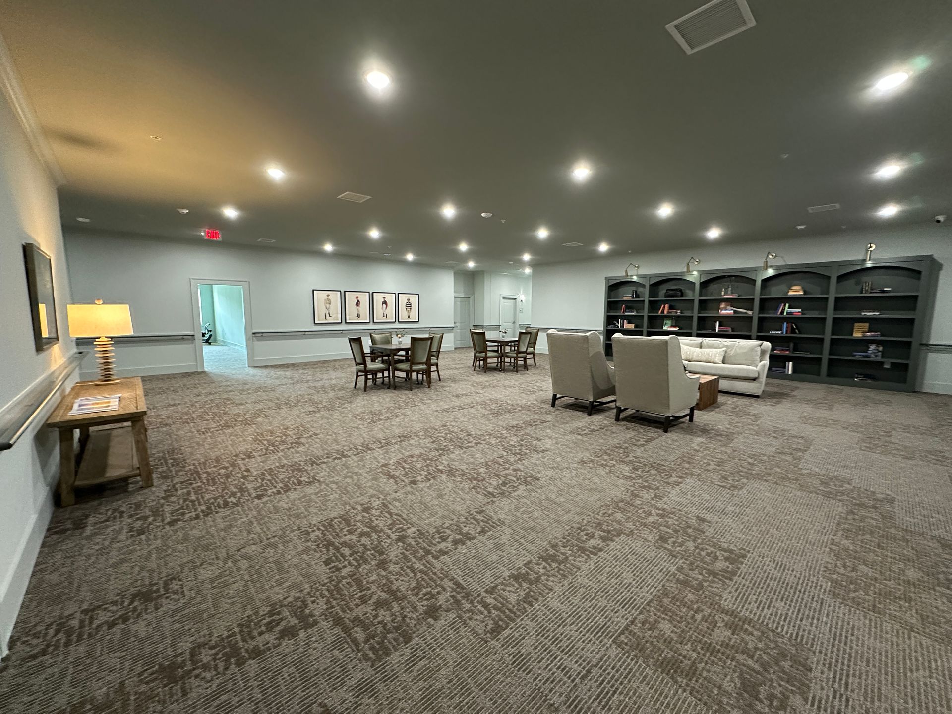 Spacious gray lounge with carpet, ceiling lights, tables, chairs, and wall shelves.