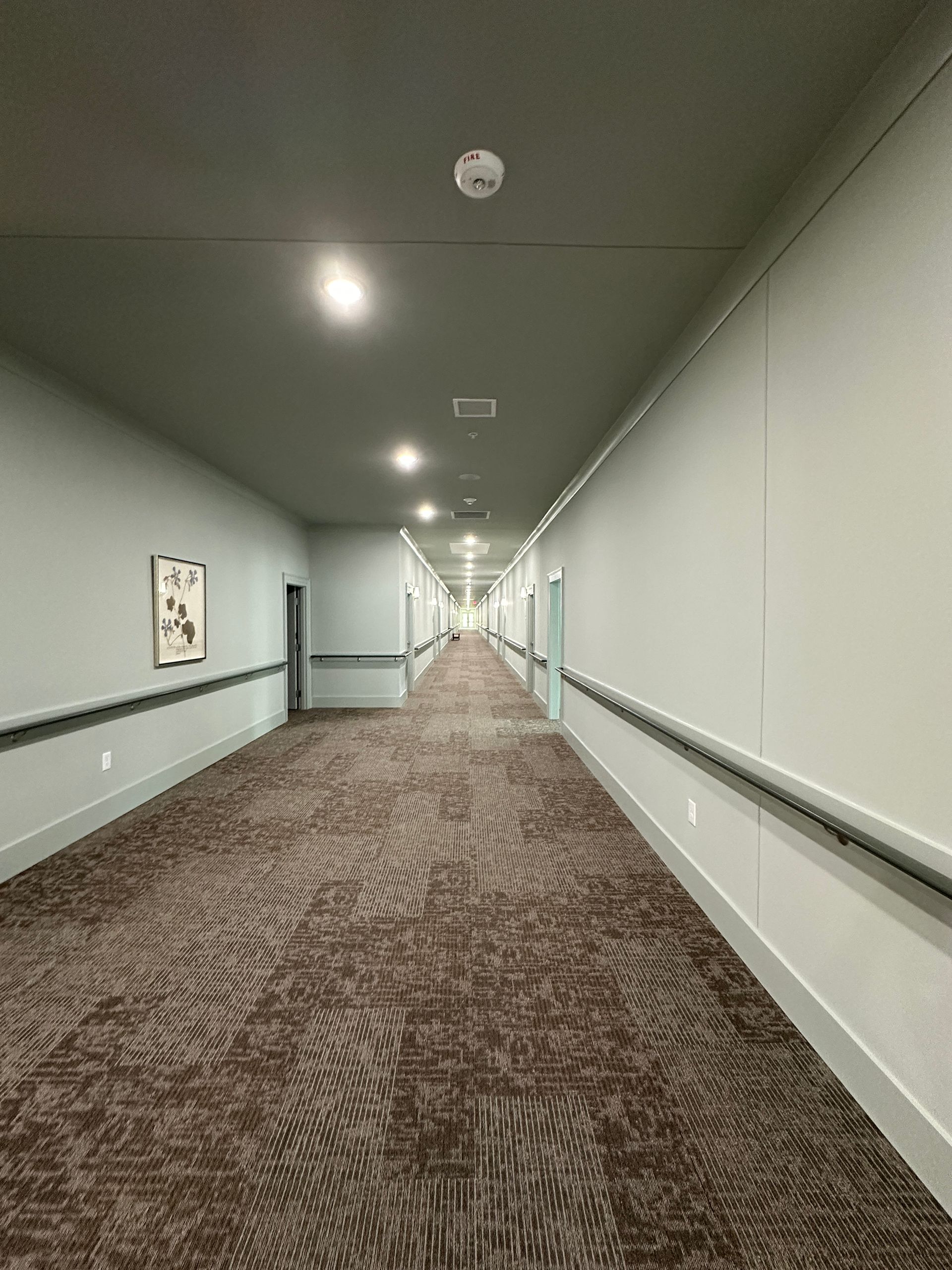 Long hotel hallway with patterned carpet, gray walls, handrails, and recessed ceiling lights