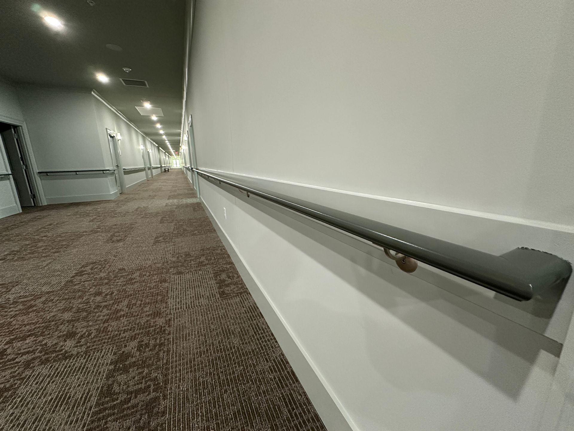Long hallway with white walls, patterned carpet, and a dark handrail along the right side