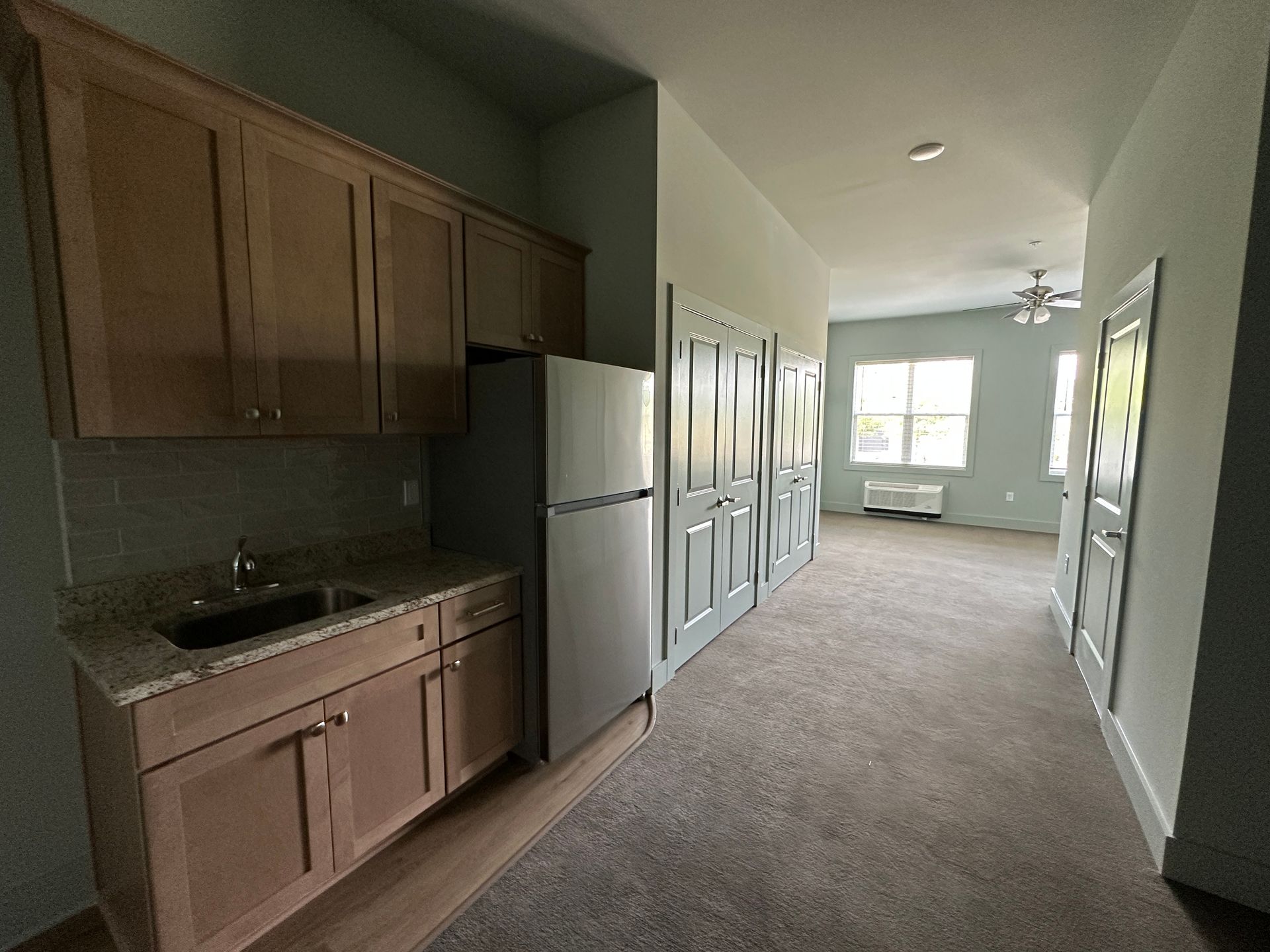 Empty apartment kitchen and hallway with light wood cabinets, white fridge, and carpeted floor leading to bright windows