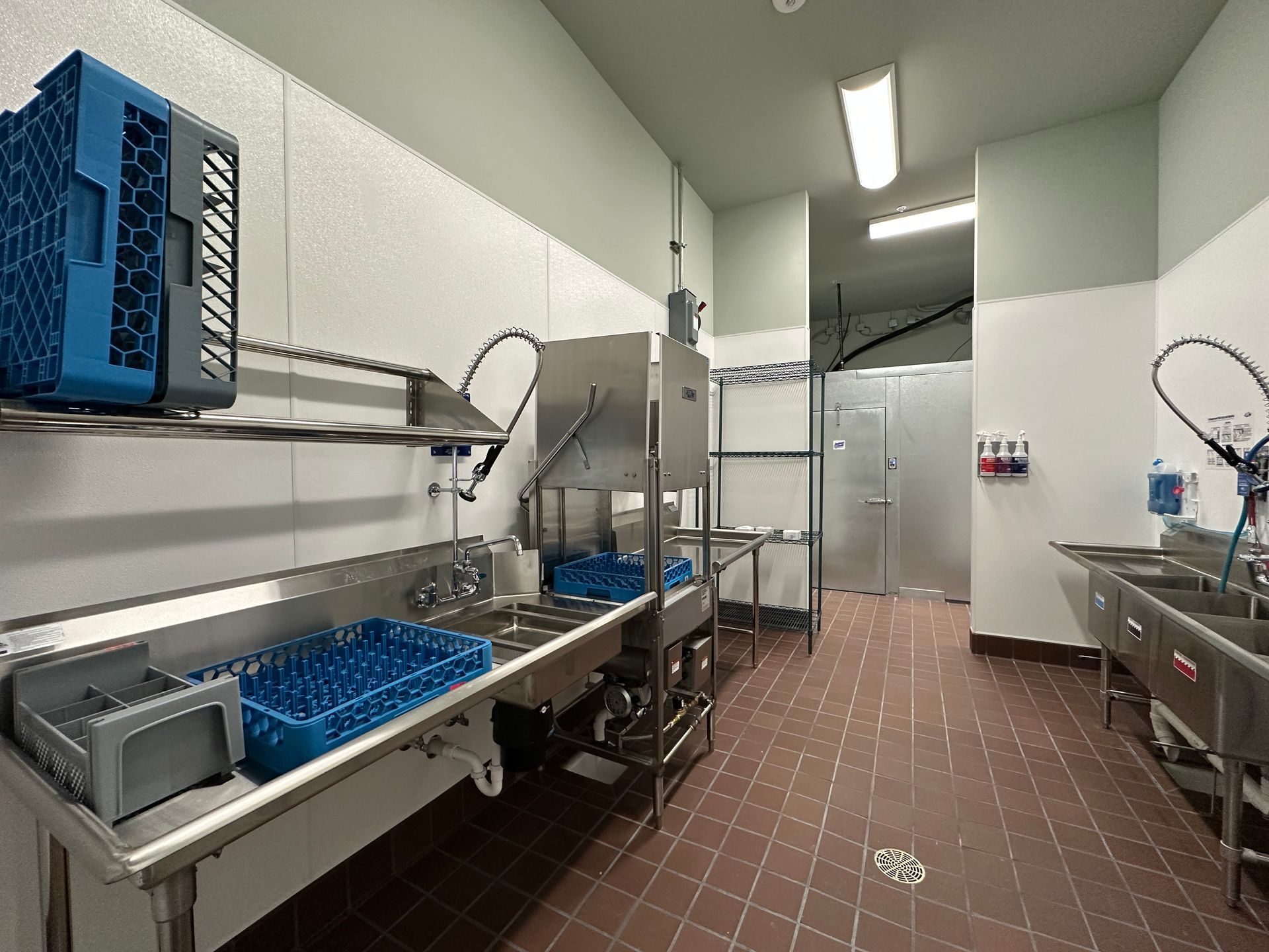 Commercial kitchen dishwashing area with stainless steel sinks, blue racks, and tiled floor.