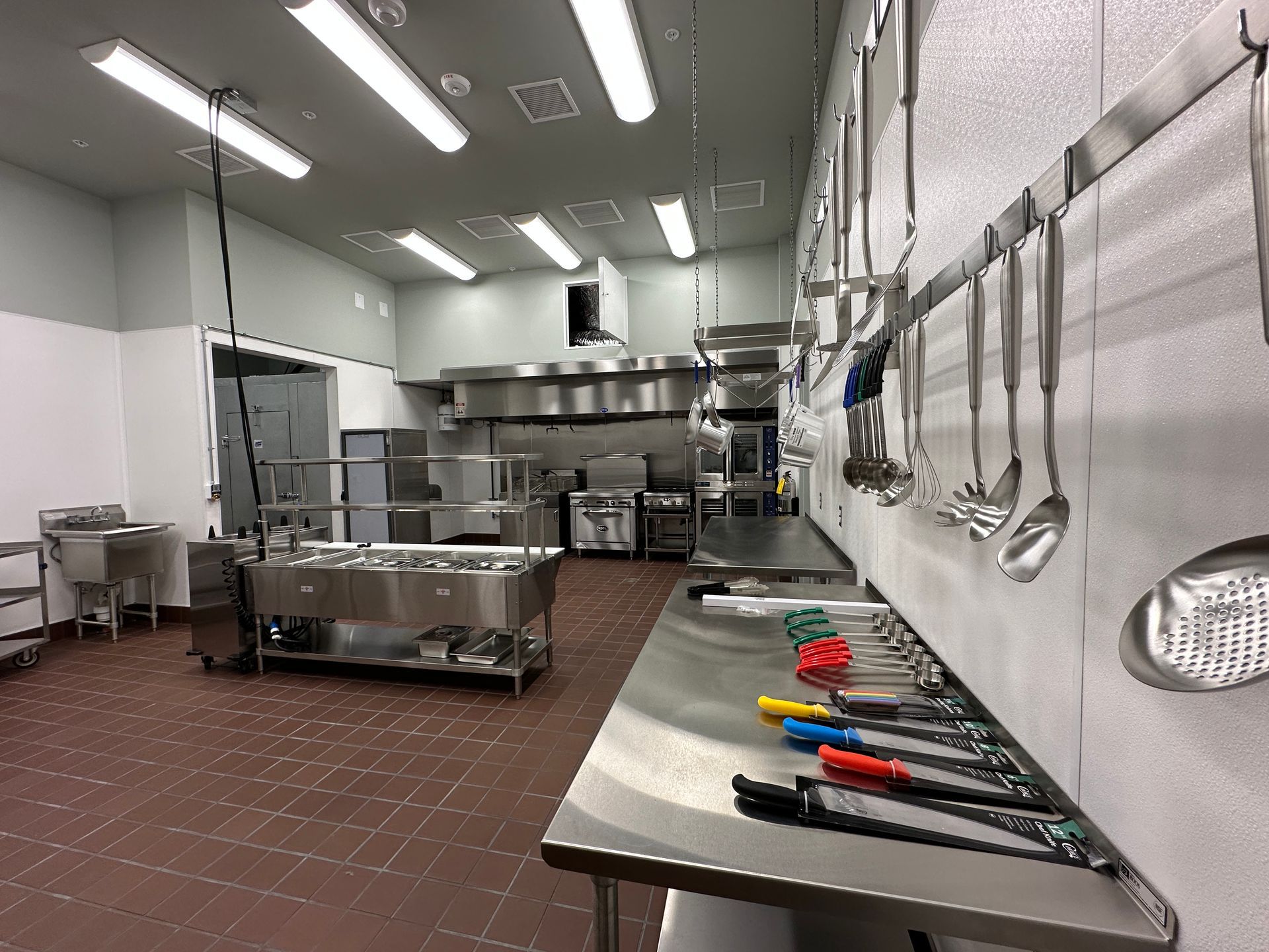 Commercial kitchen with stainless steel counters, hanging utensils, and prep tools on a worktable.