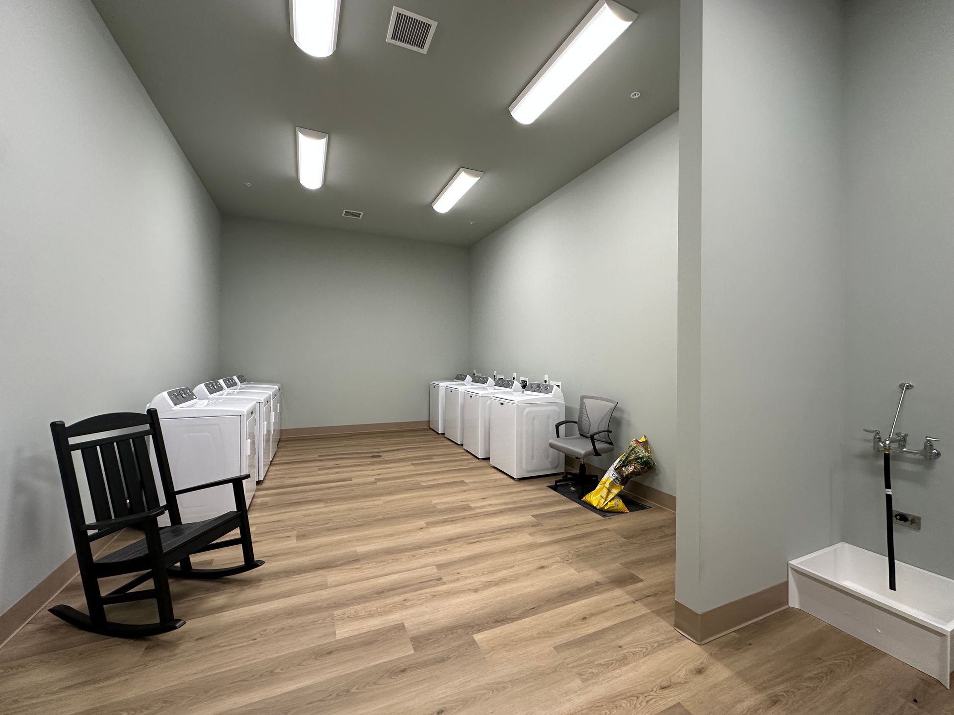 Empty laundry room with washing machines, a black chair, and a mop sink on light wood flooring.