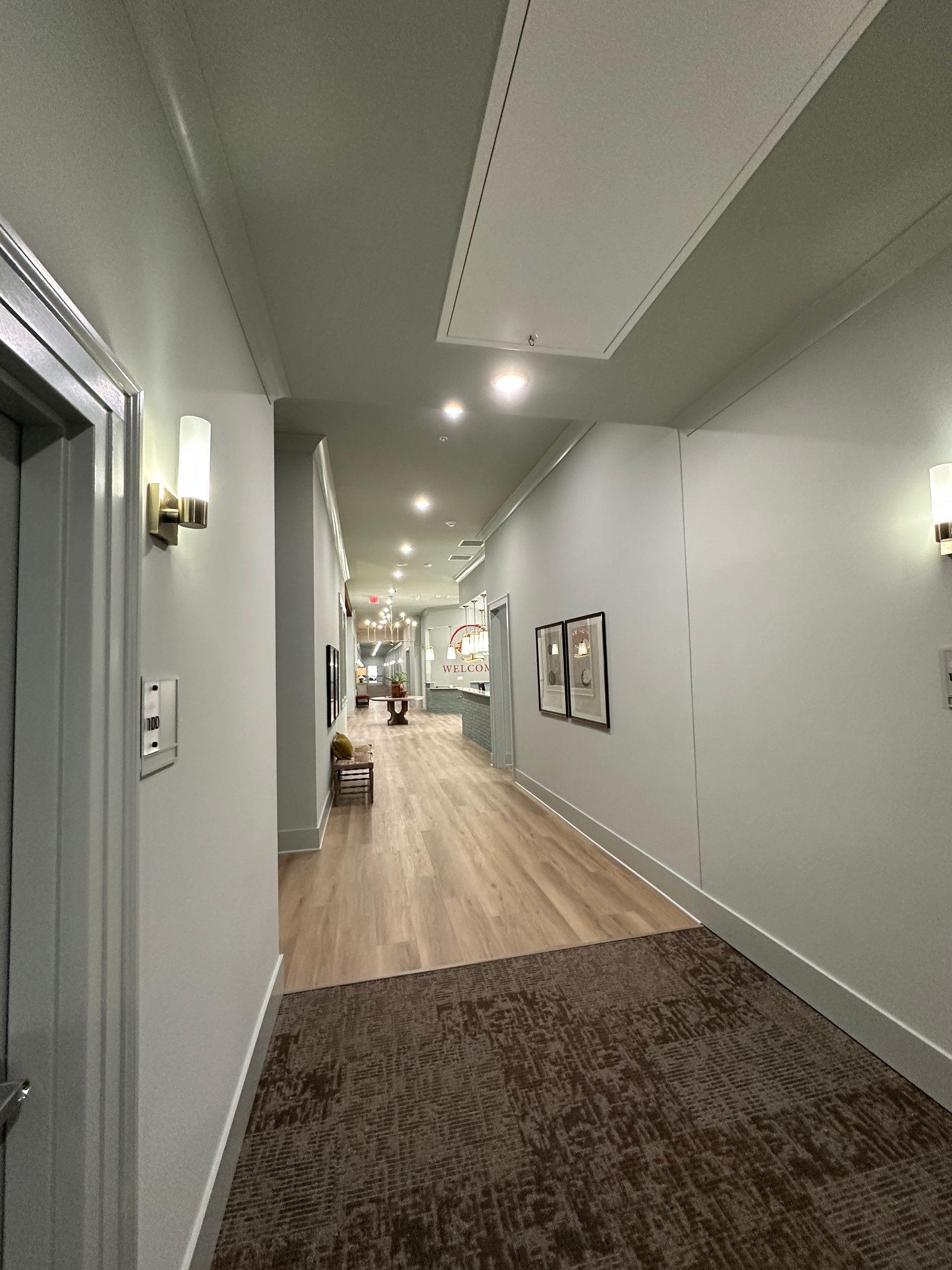 Modern hallway with gray walls, wall sconces, and light wood flooring leading to a distant seating area.