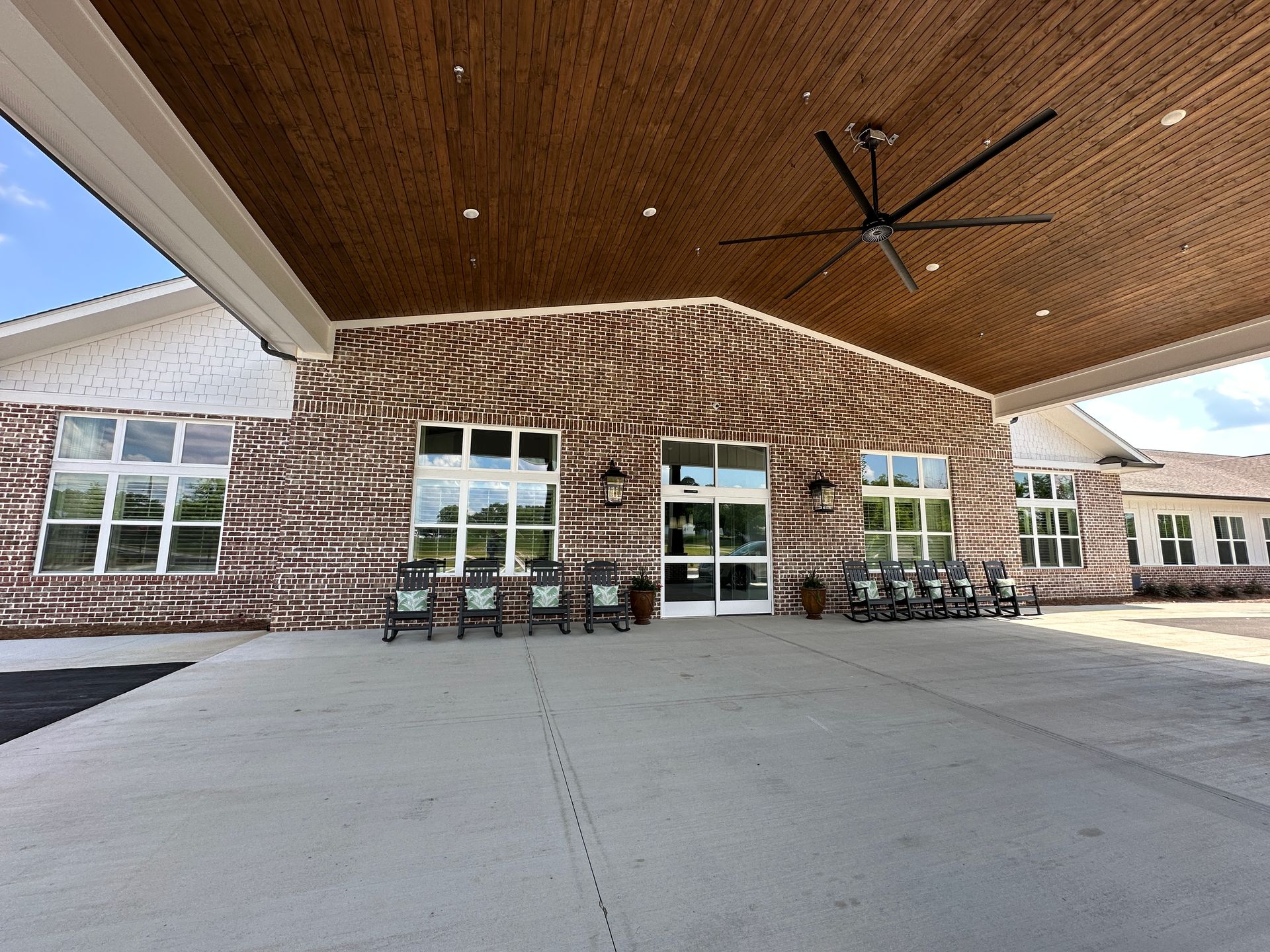 Covered brick building entrance with large windows, benches, and wide concrete patio under a wooden overhang