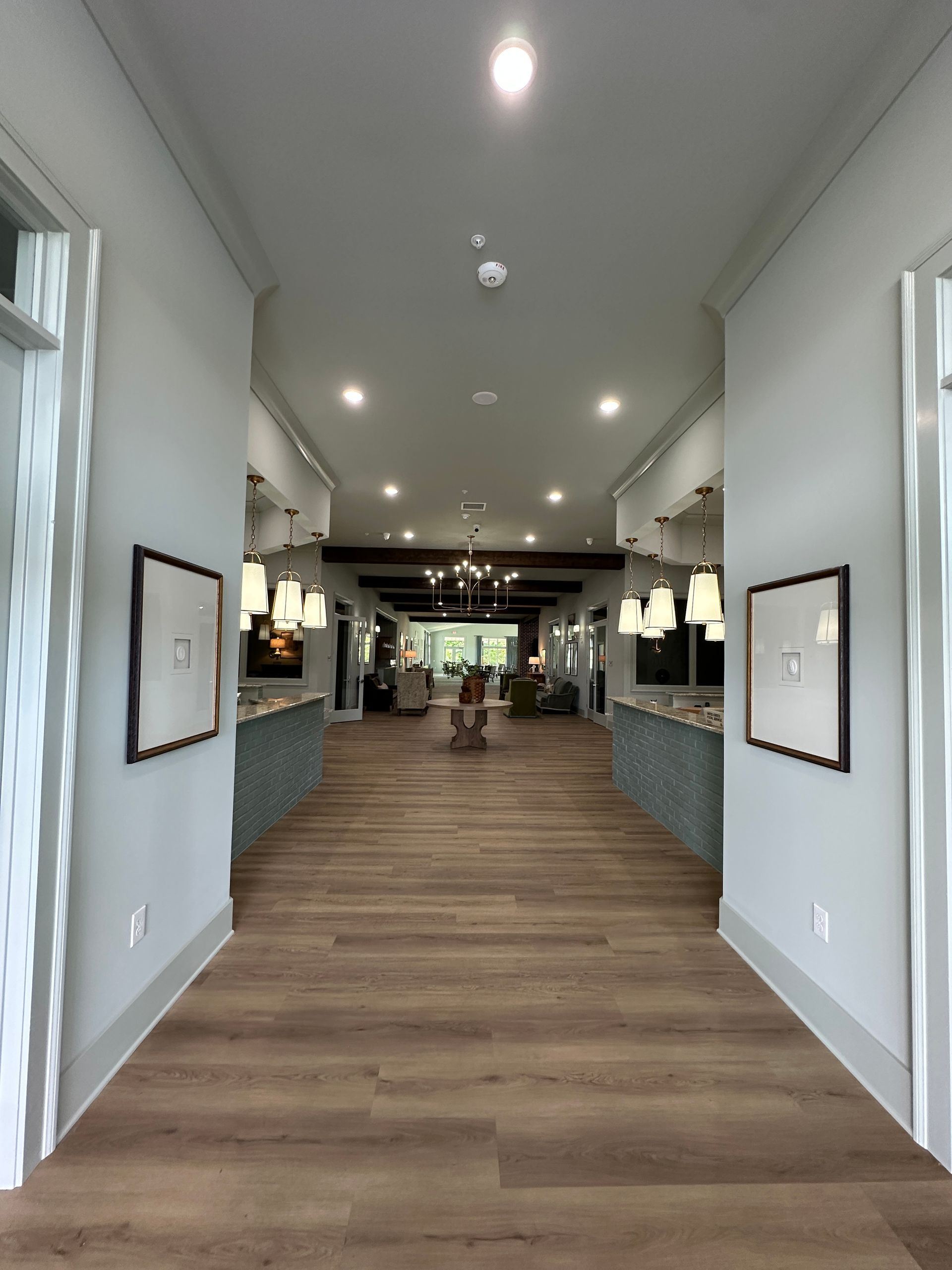Bright hallway with wood floors, white walls, recessed lights, and seating areas leading to a lobby.