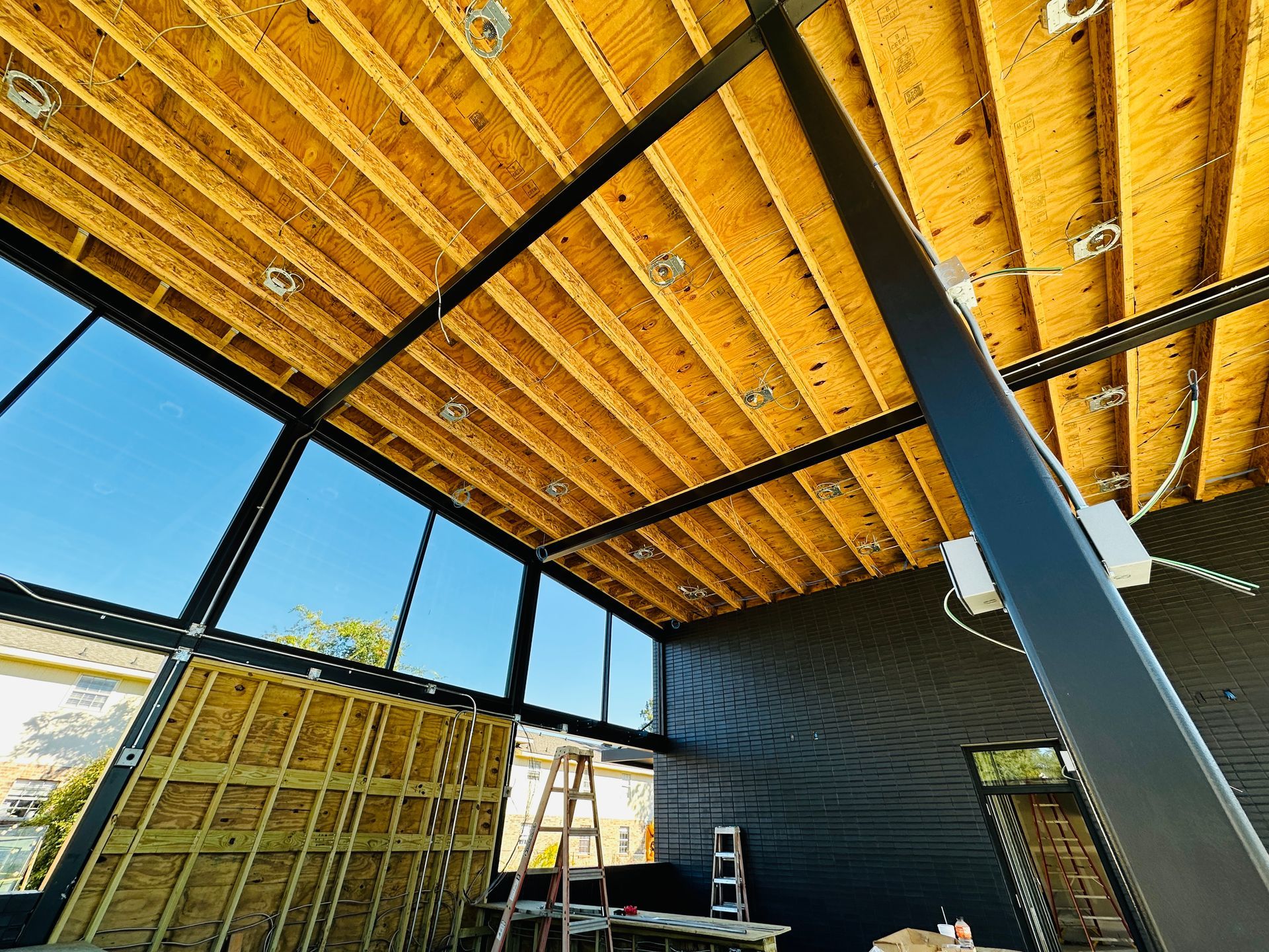 Unfinished interior with exposed wood ceiling beams, large windows, and ladders against a dark wall.