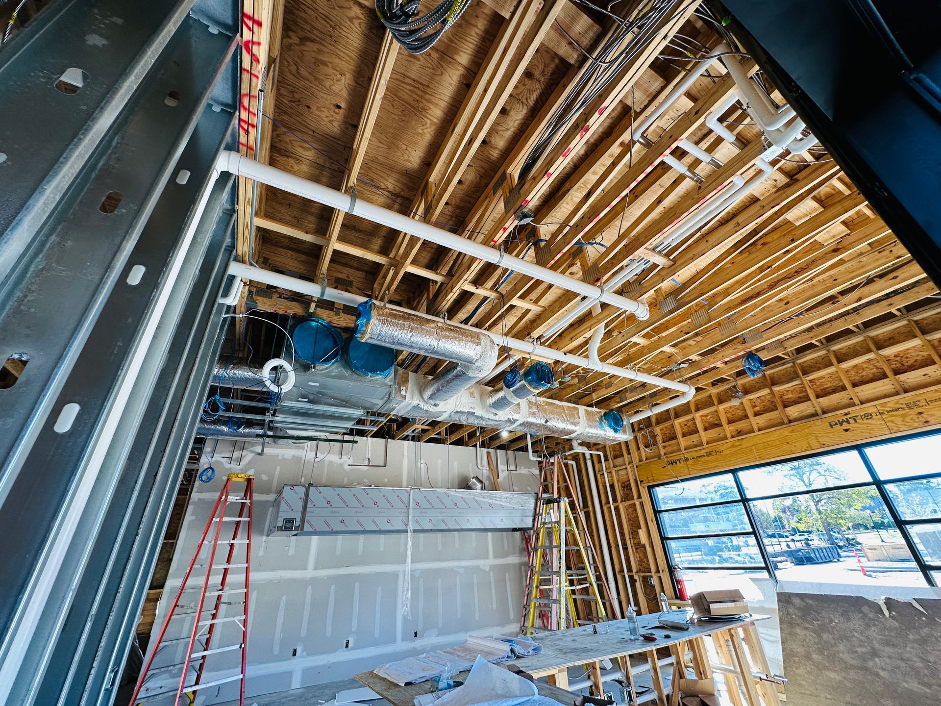 Construction site interior with exposed wood ceiling, steel framing, ladder, and hanging ductwork and lights
