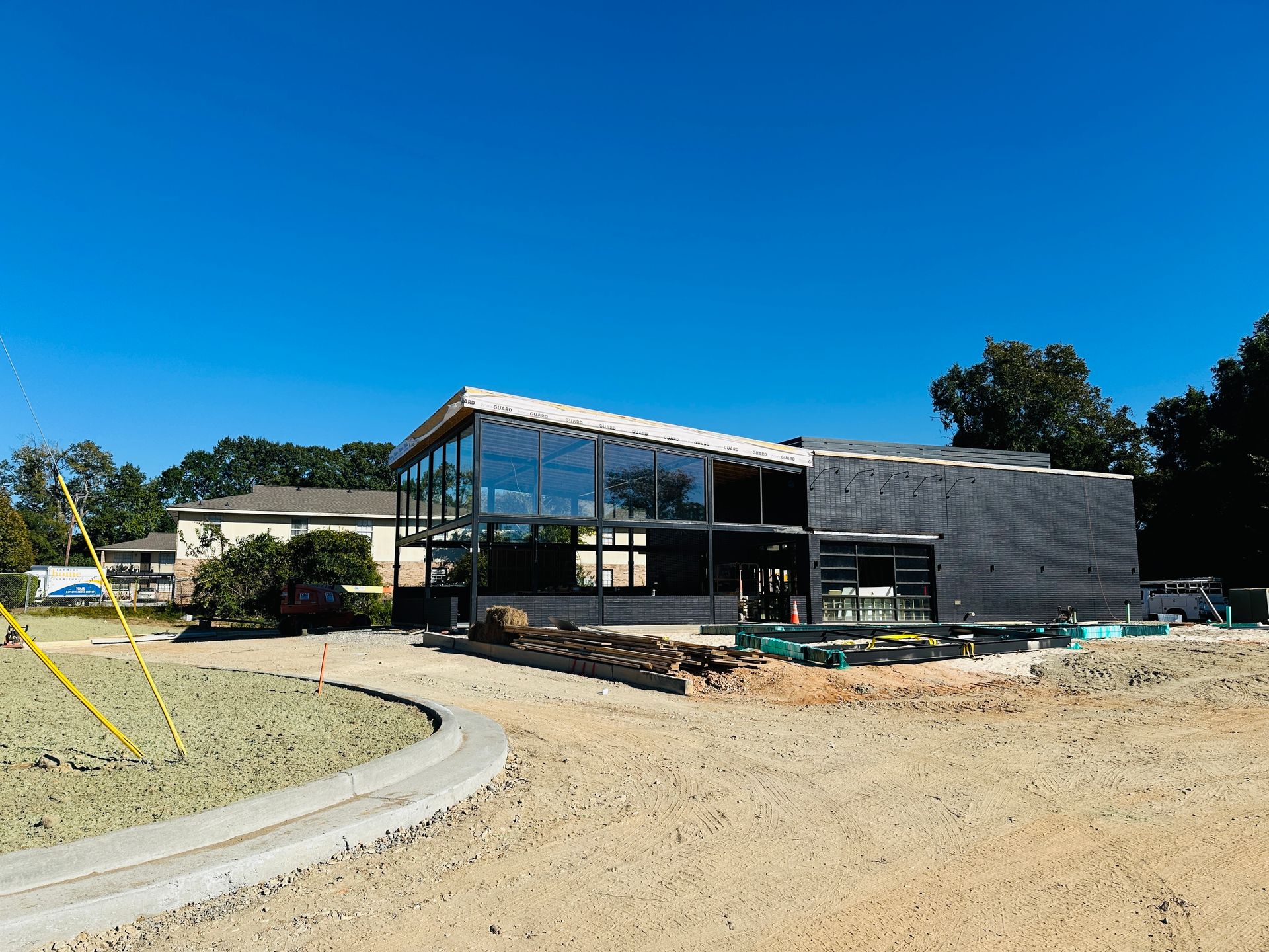 Modern building under construction with glass facade, gray siding, and dirt foreground under a clear blue sky