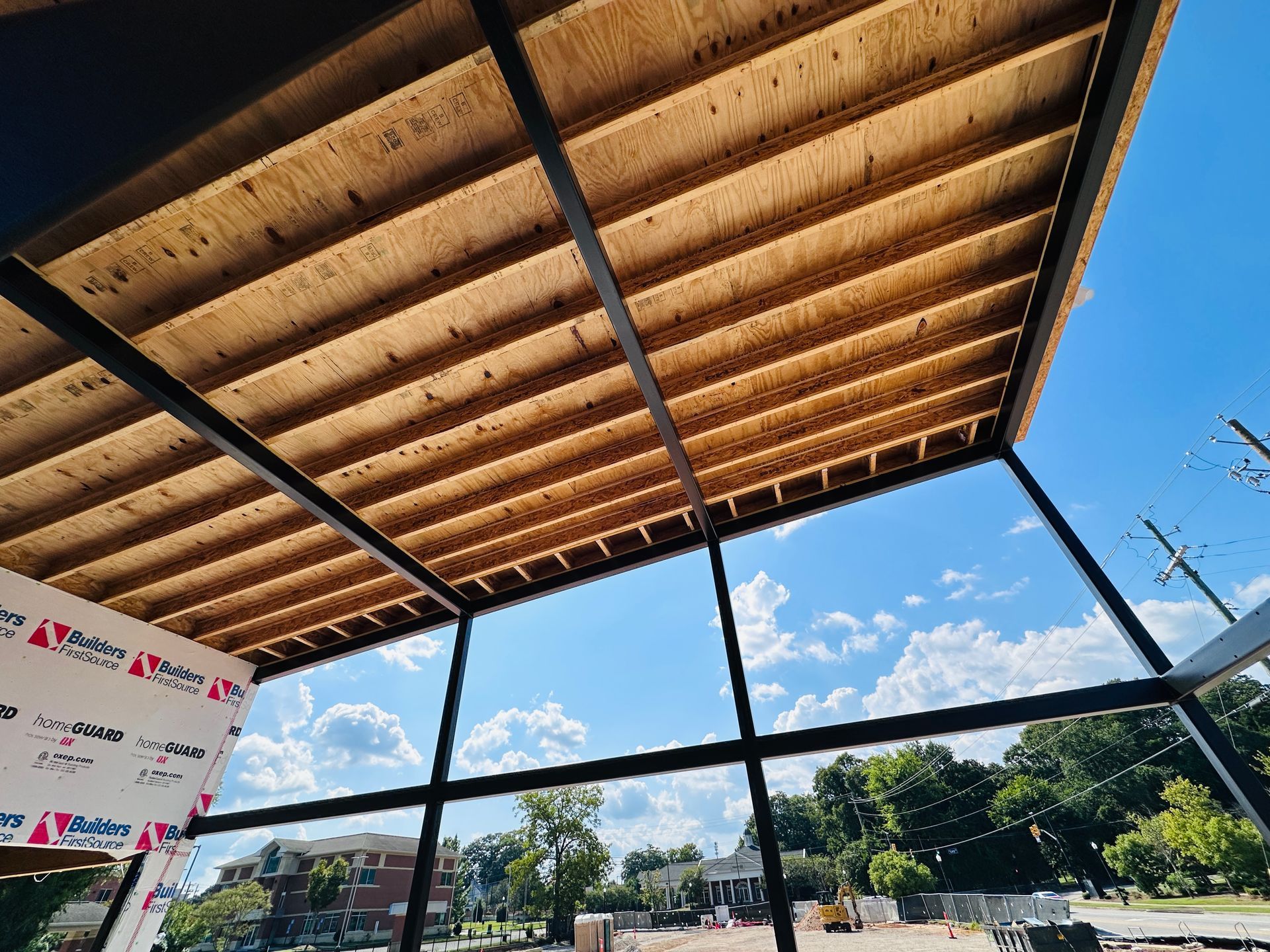 Open-air pavilion with wooden roof, black beams, and a bright blue sky with clouds beyond.