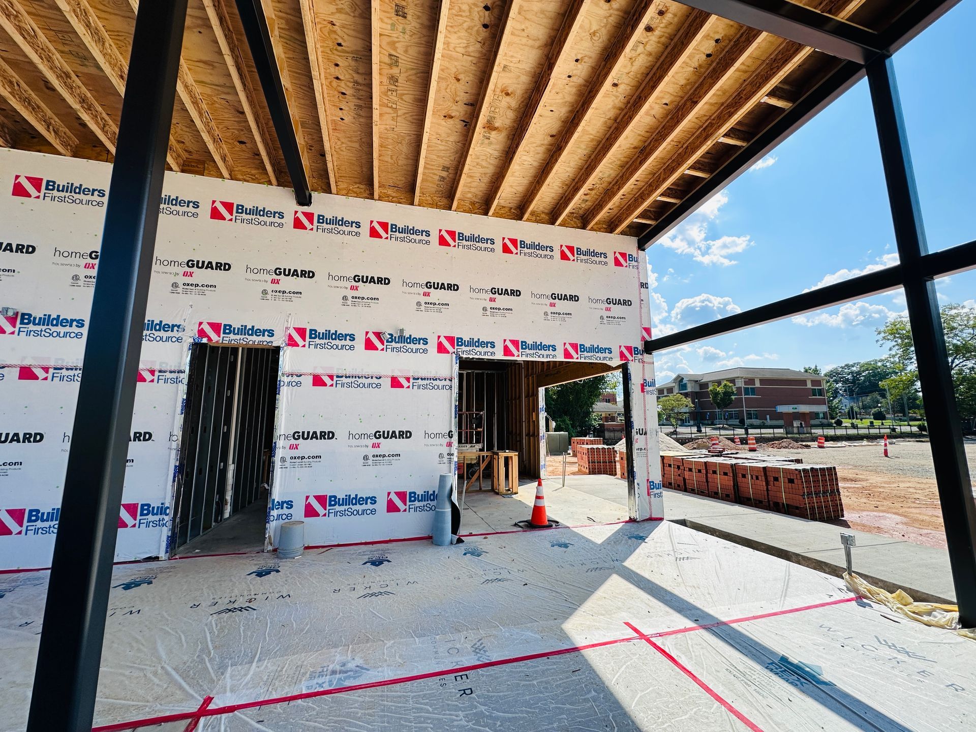 Construction site interior with exposed wood ceiling, black window frames, and wrapped walls near an open doorway.