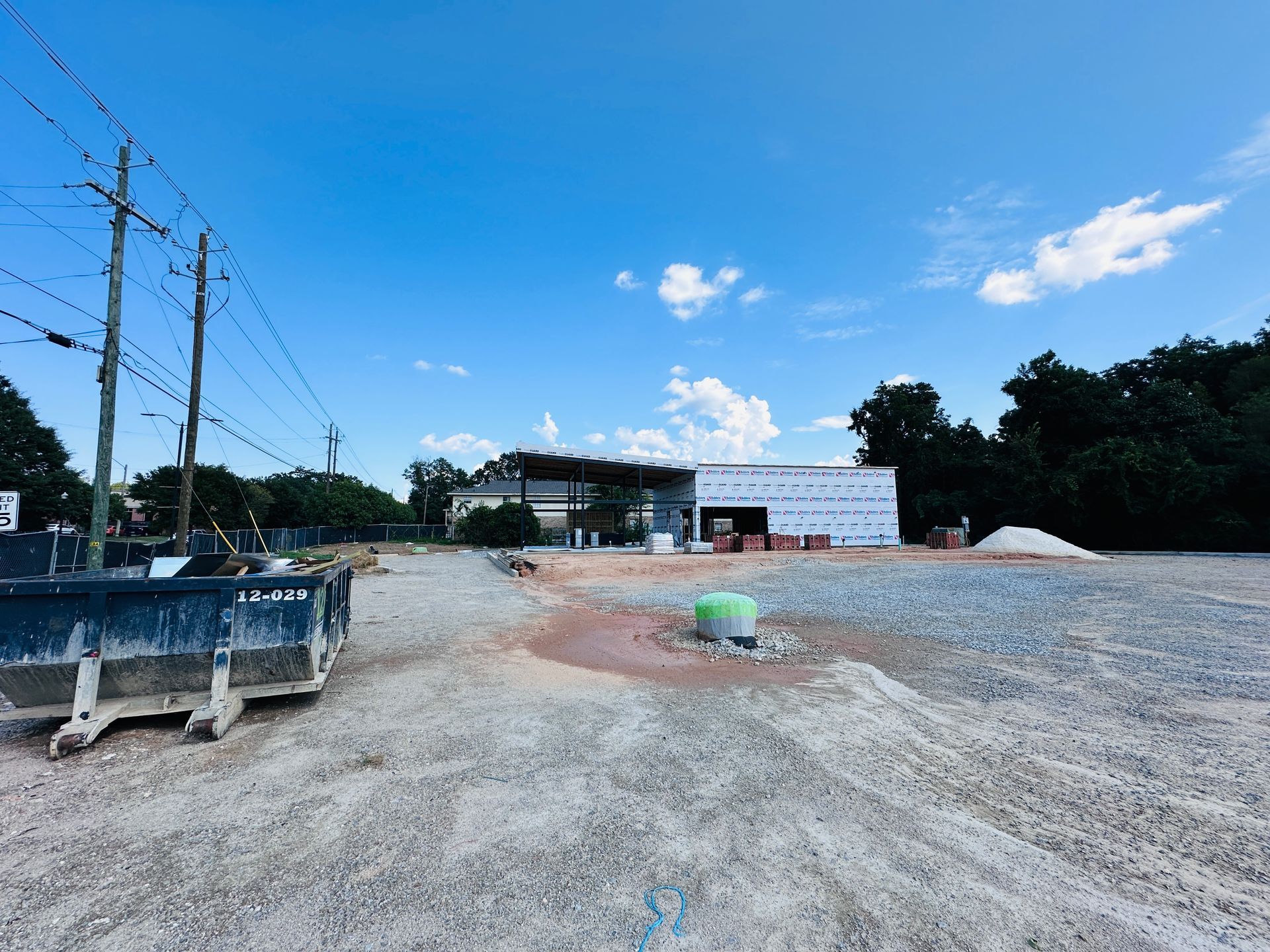 Wide gravel construction site with utility poles, a white building, and trees under a blue sky