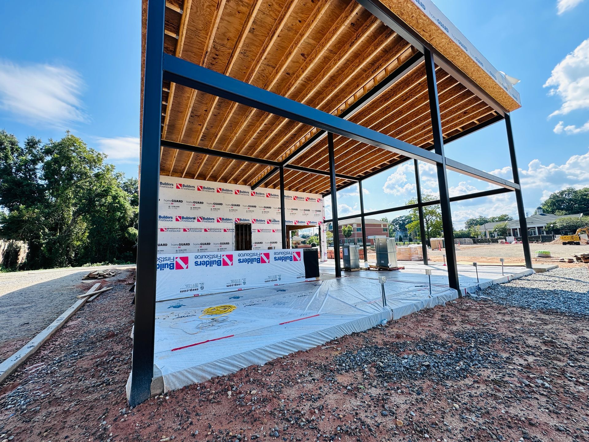Open-air pavilion under construction with black steel posts, wood roof, and white tarp flooring