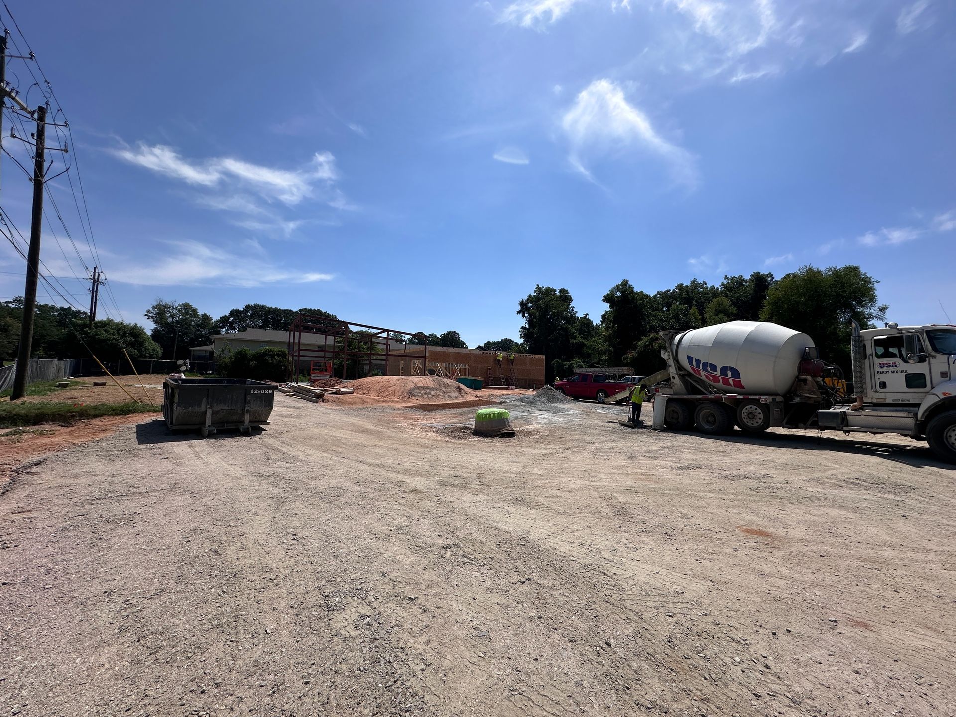 Gravel lot with a cement mixer truck, construction materials, and a partly cleared worksite under a blue sky