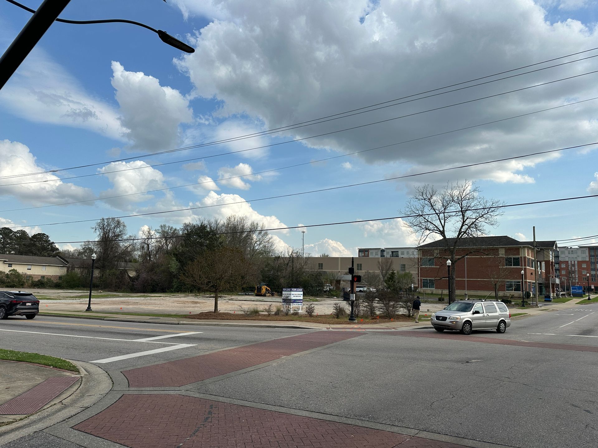 A street-level view of a vacant, dirt-covered lot at a sunny urban intersection with a few cars and trees in the distance.