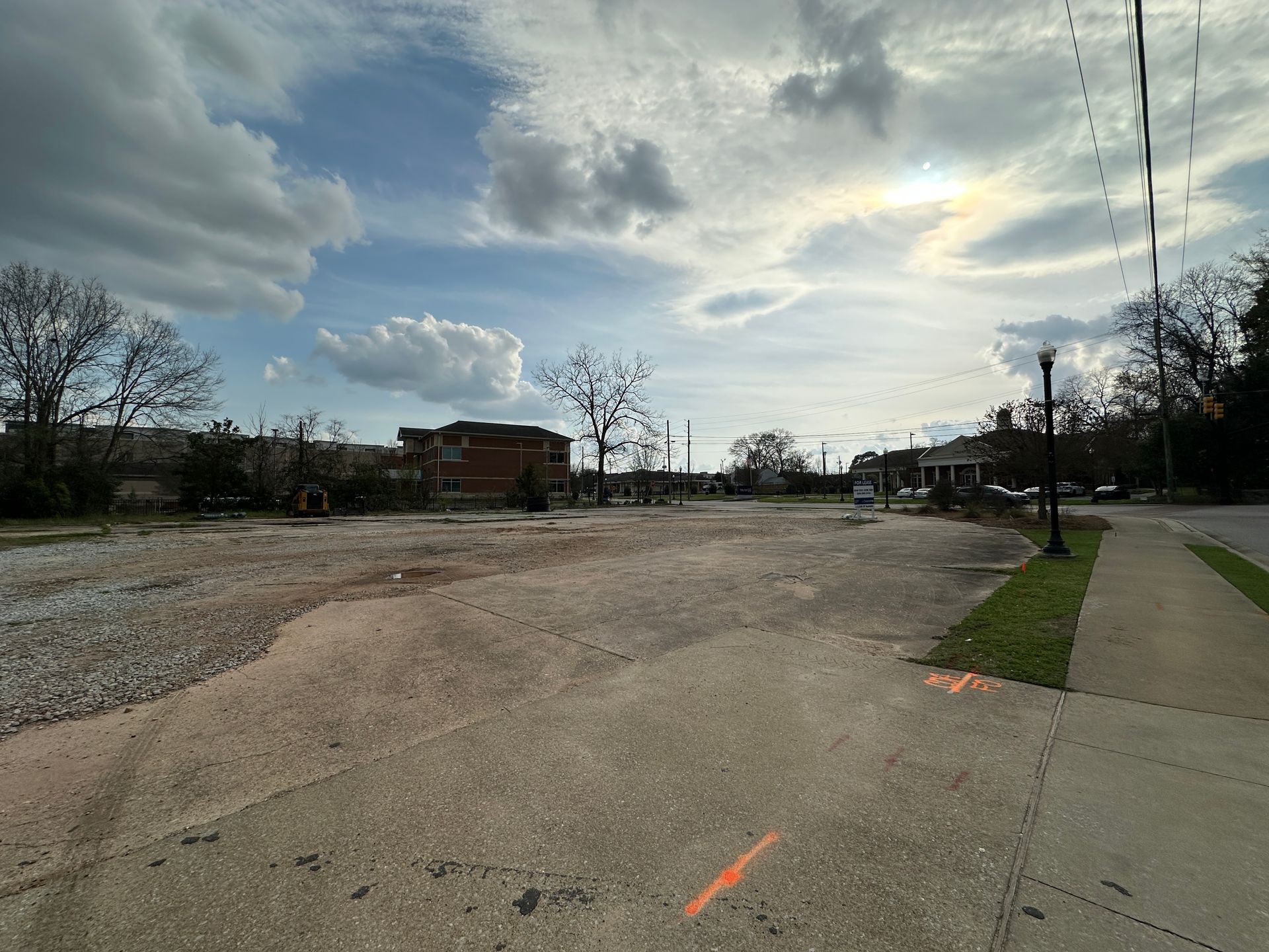 A wide, empty, gravel-covered lot under a cloudy sky, adjacent to a sidewalk and a street with distant buildings.