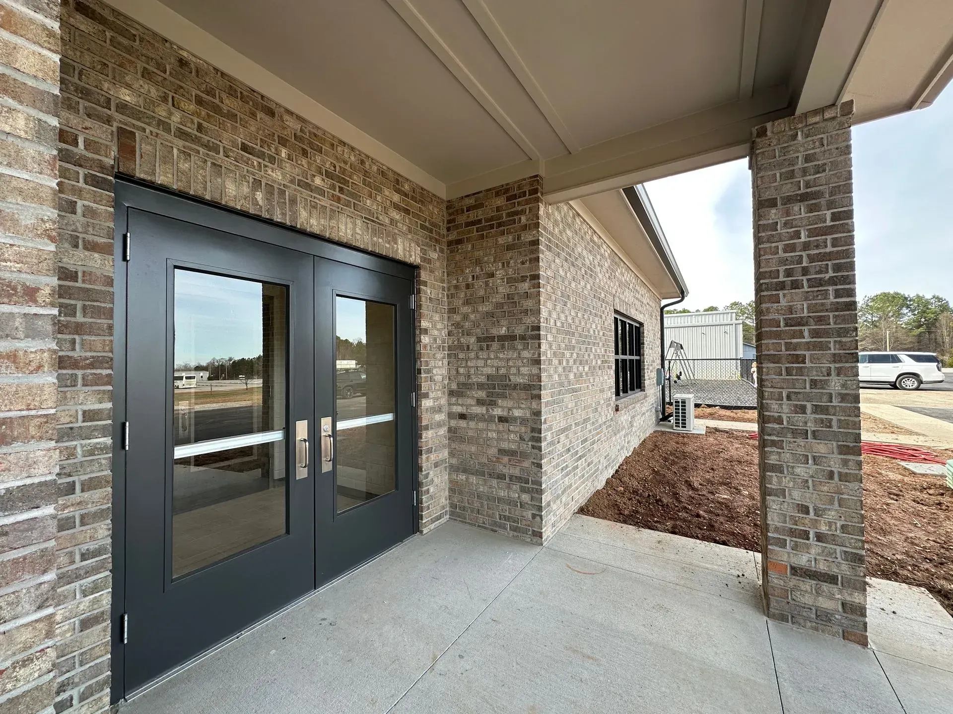 Covered brick entryway with dark double glass doors and a concrete sidewalk leading to a building