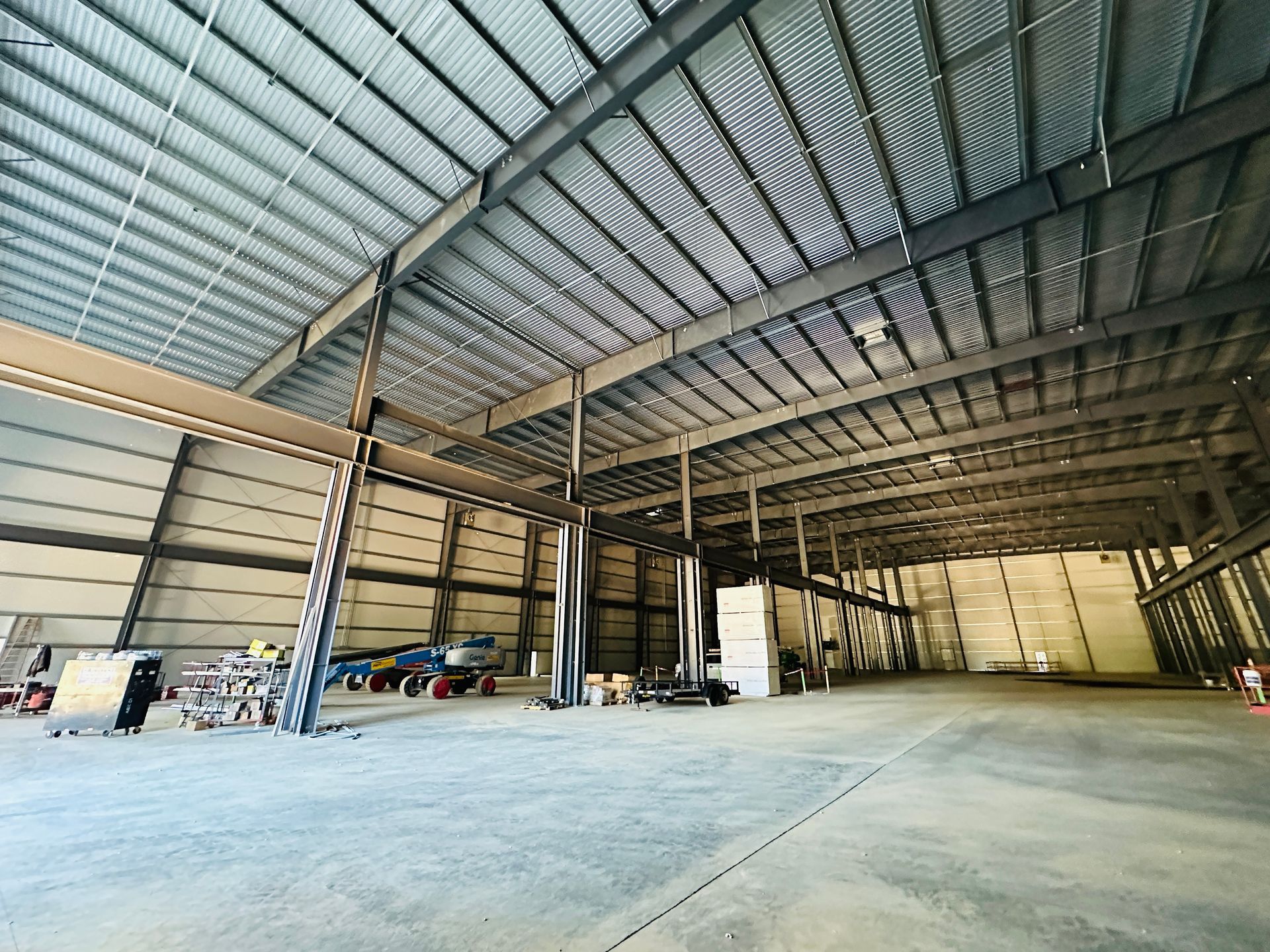 Empty industrial warehouse with high metal roof, concrete floor, and stacked equipment along the walls