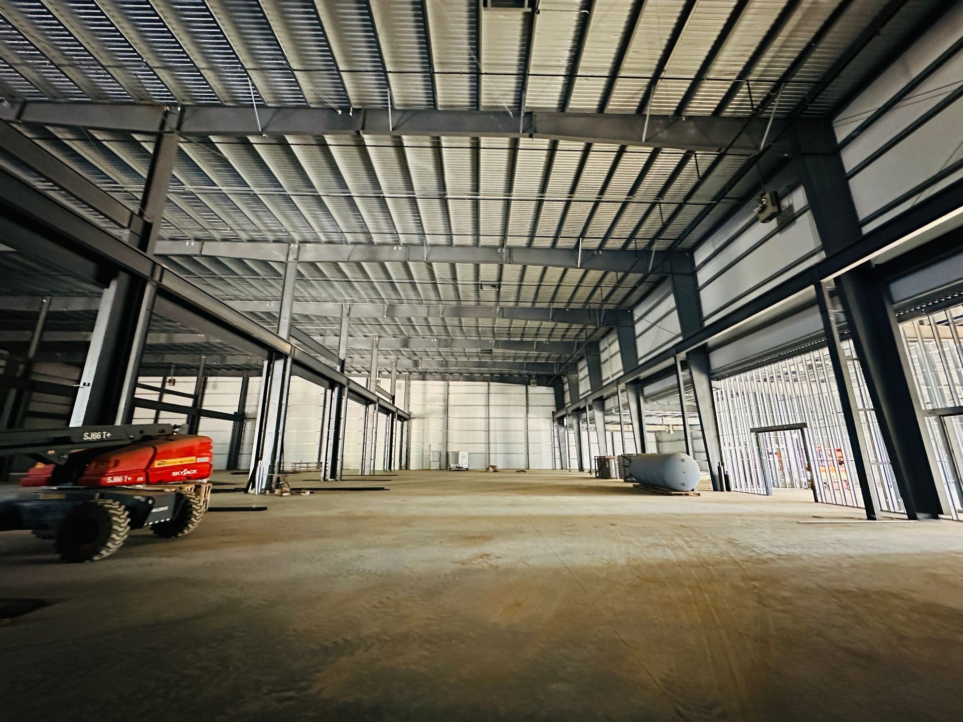 Interior of a large unfinished warehouse with steel beams, concrete floor, and a red lift on the left