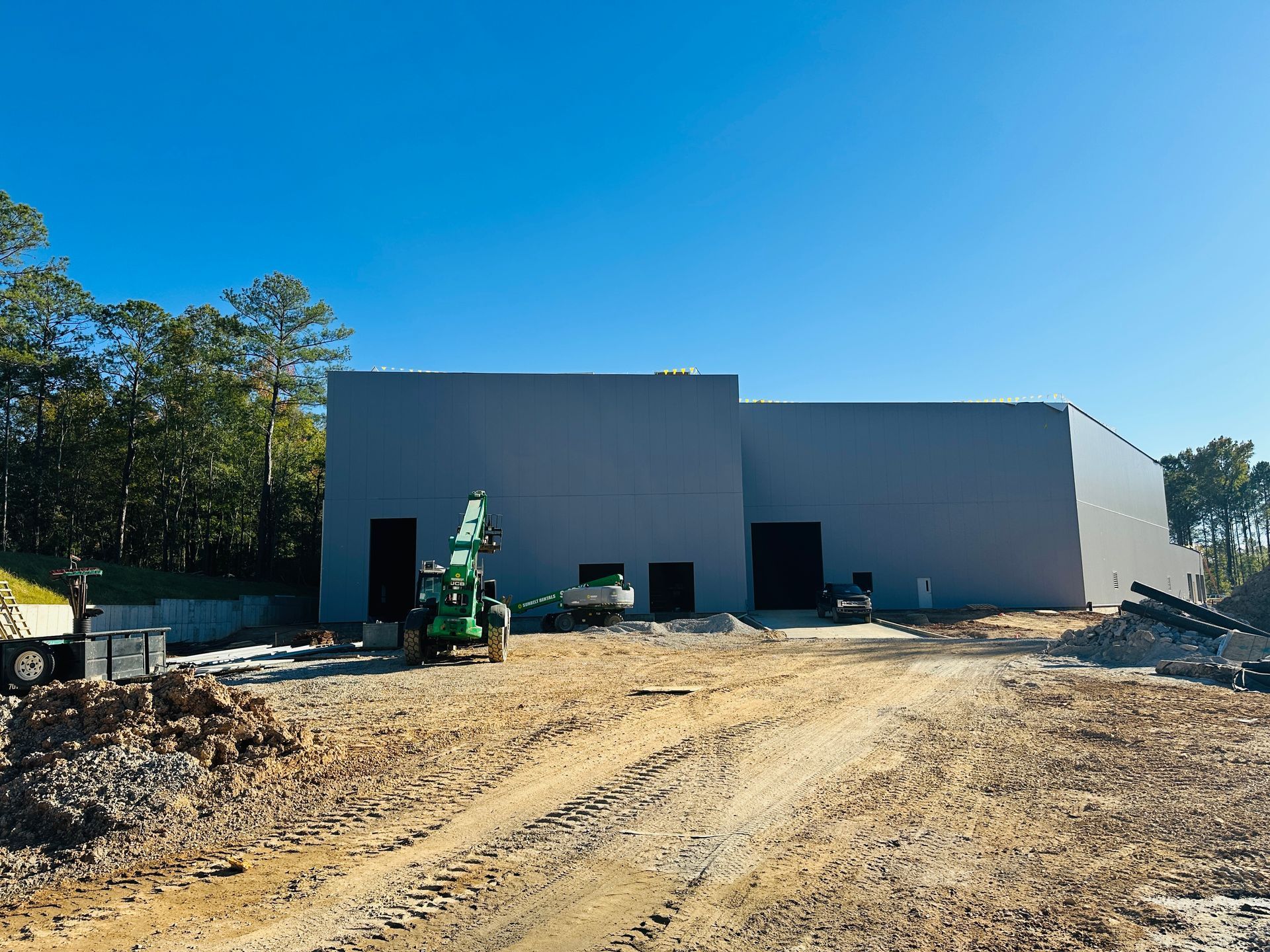 Construction site with a large gray building, dirt road, and a tractor under a clear blue sky