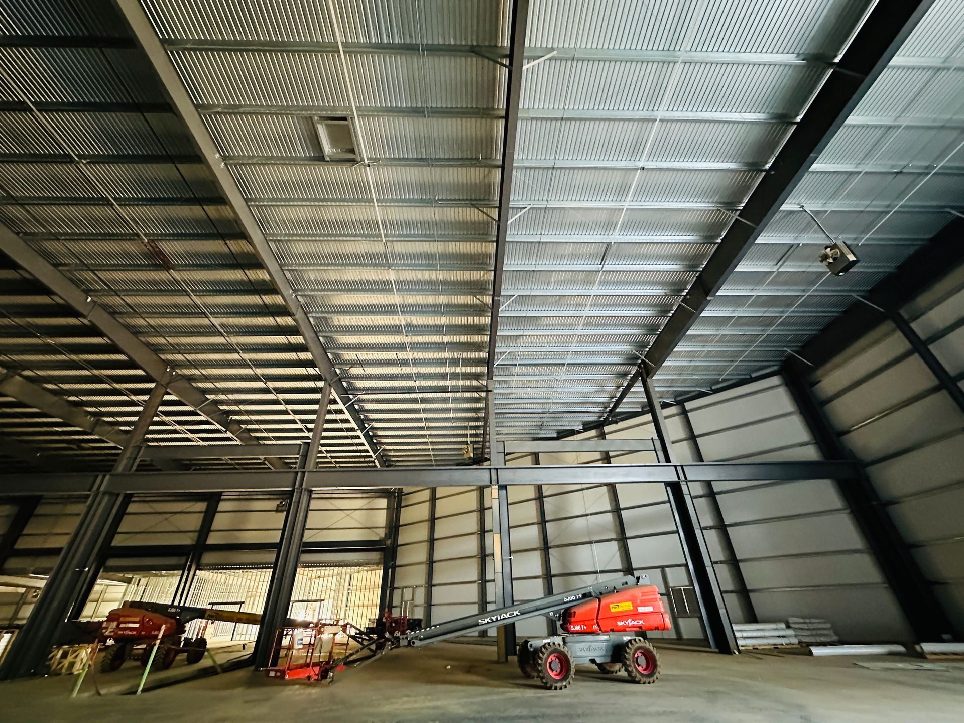 Large empty metal warehouse interior with high ceiling, steel beams, and a red scissor lift near the wall