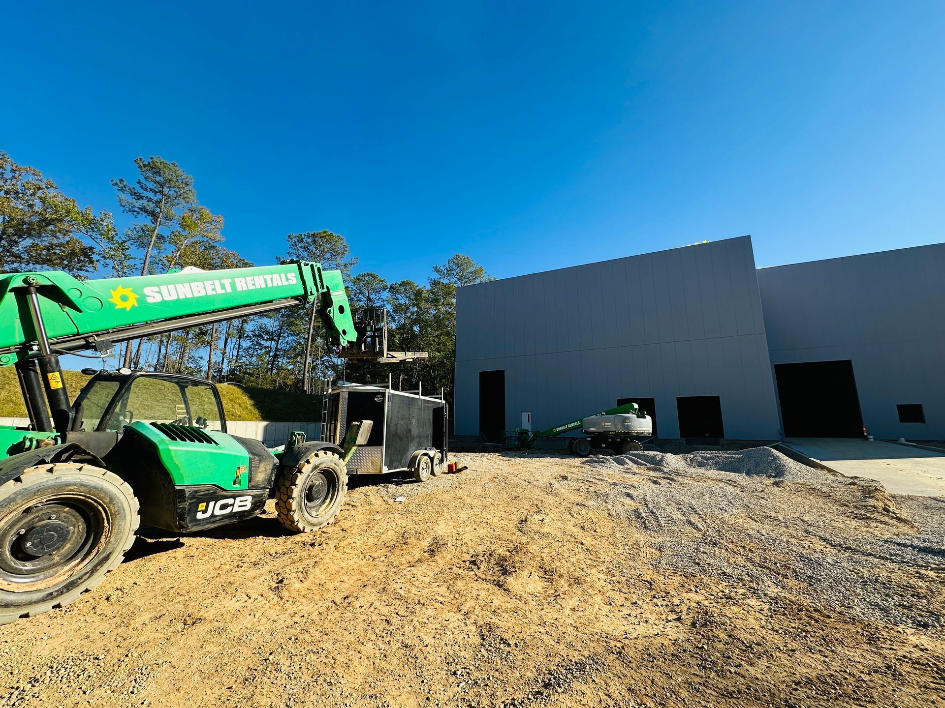 Green construction lift beside a gray building under construction on a gravel lot, under a bright blue sky