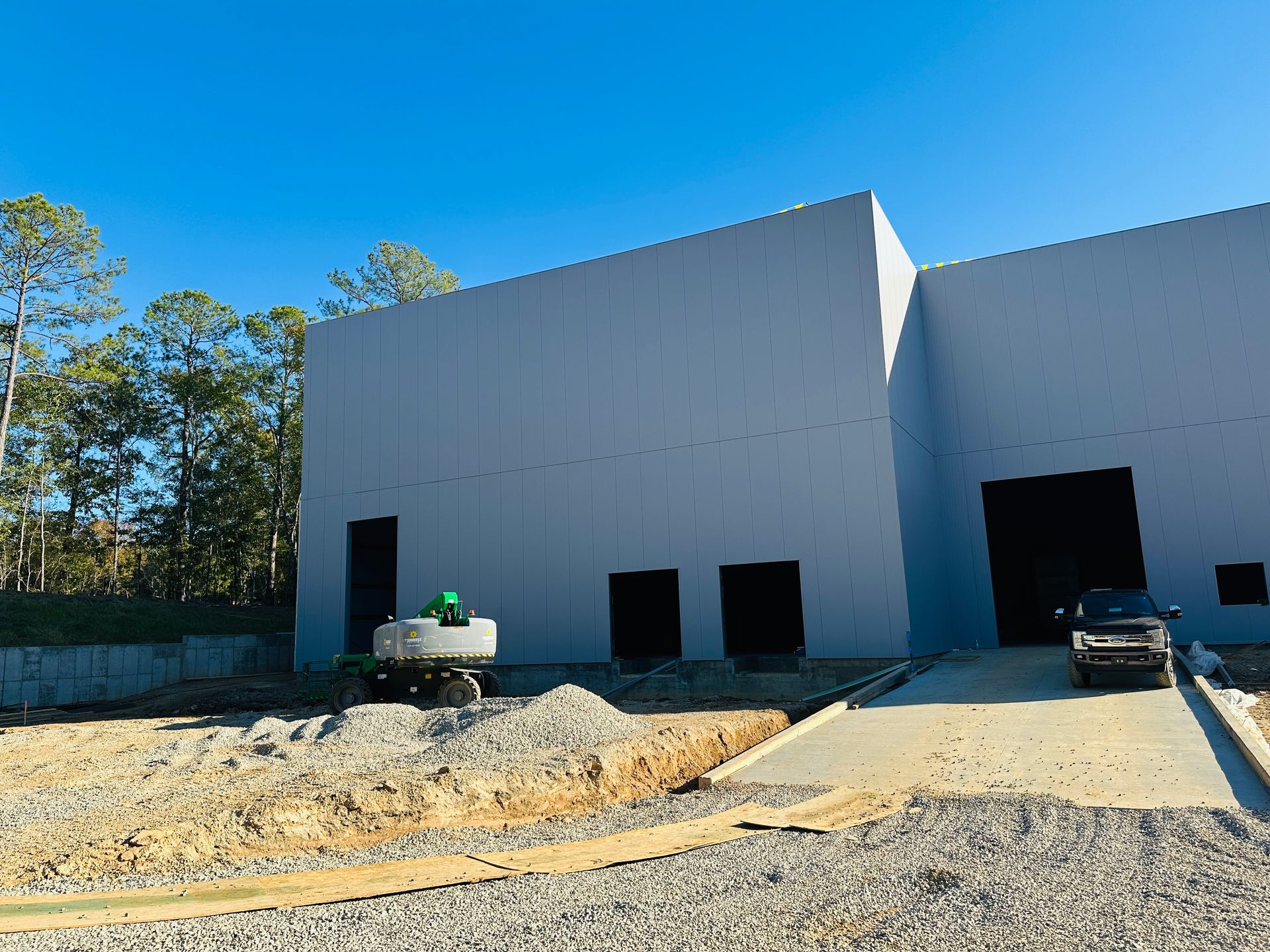 Partially built gray warehouse with open doorways, dirt lot, and construction equipment under a blue sky