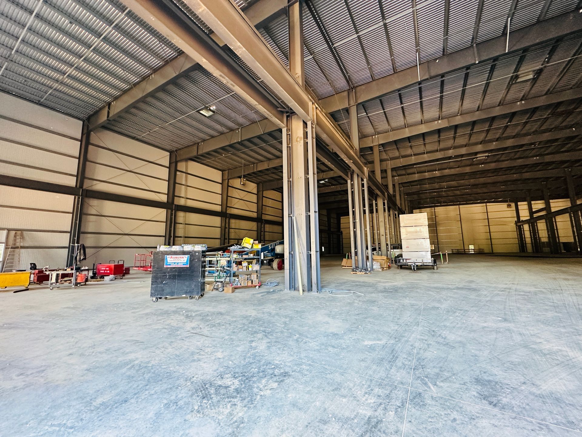 Empty warehouse interior with metal beams, equipment, and stacked pallets on a concrete floor