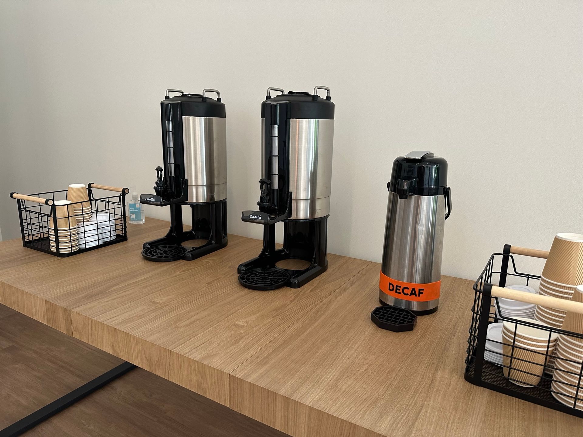 A wooden table with three coffee dispensers and a basket of cups on it.