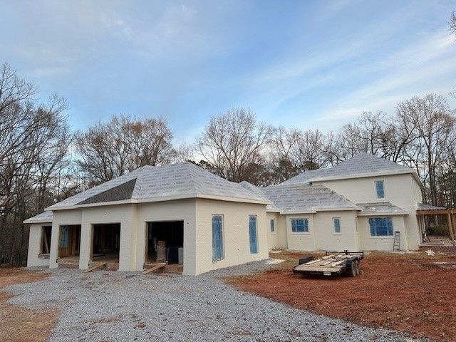 Construction of a light beige stucco house with gray roof, unfinished garage, and blue-taped windows on a gravel lot.