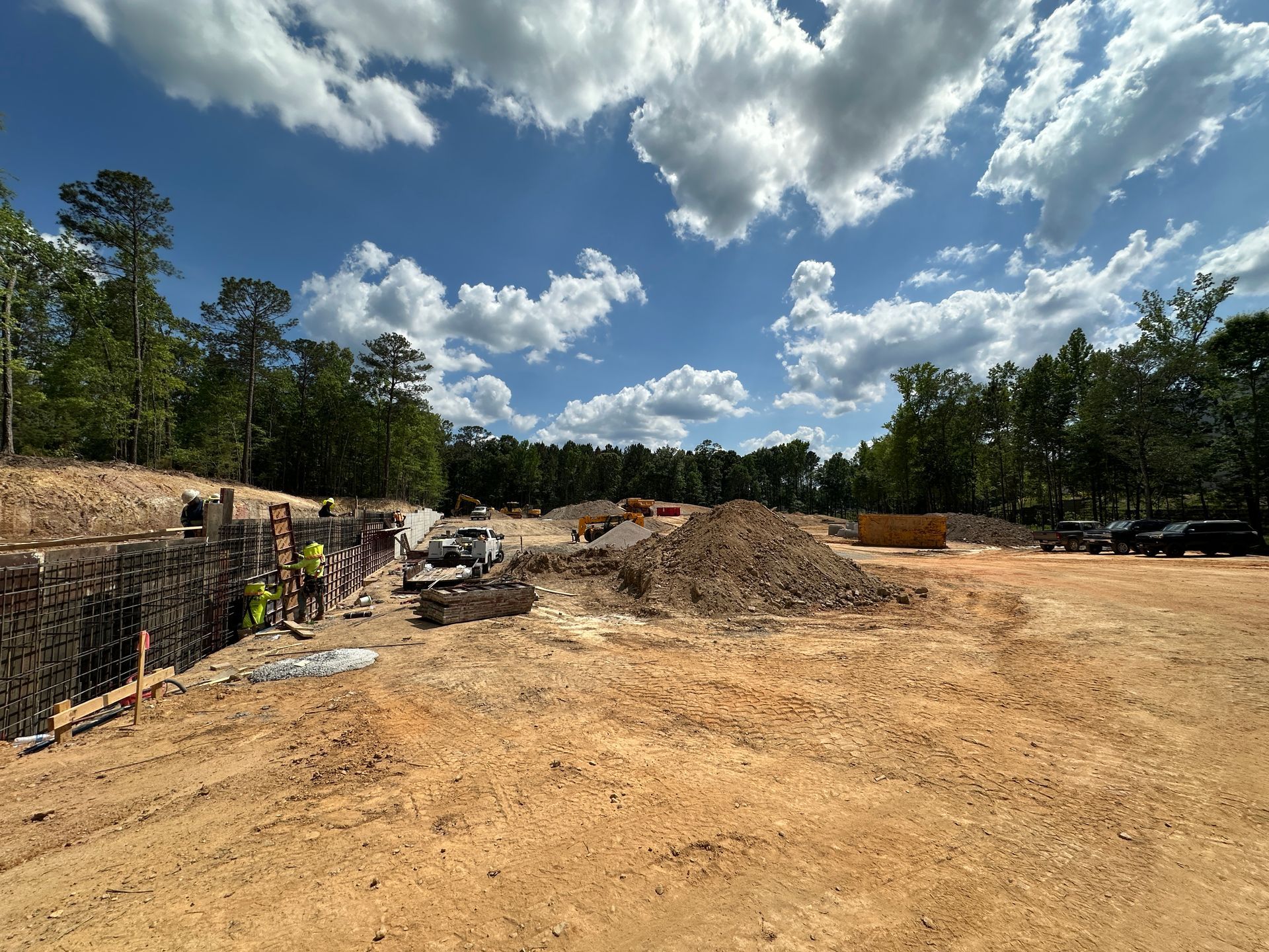 Cleared construction site with initial foundation wall, dirt piles, heavy equipment, and trees under a bright cloudy sky