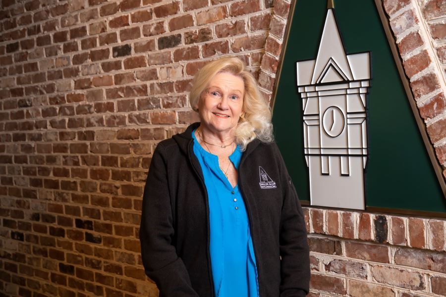 A woman in a blue shirt and black jacket is standing in front of a brick wall.