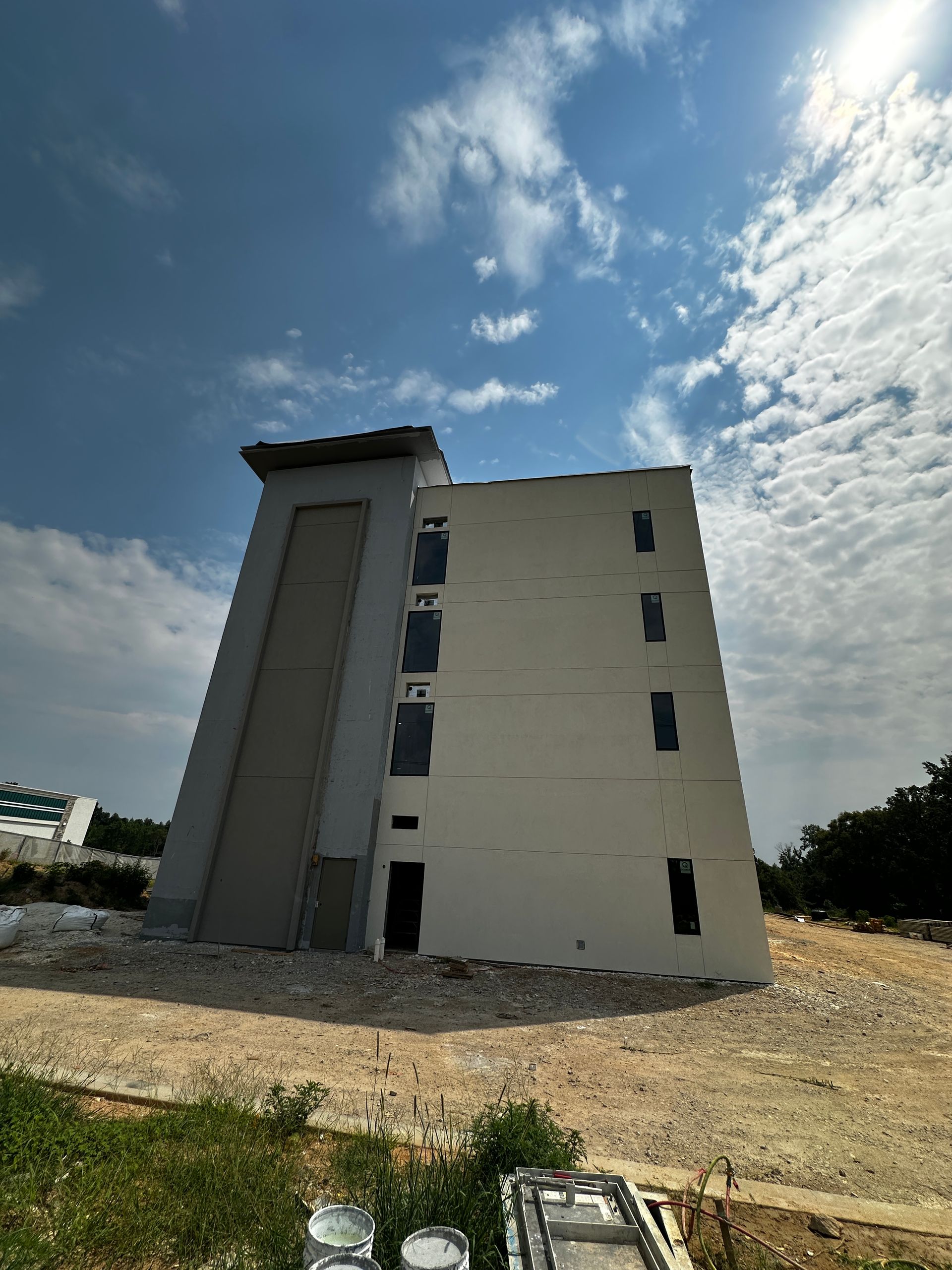 Tall beige building with a gray annex under a cloudy blue sky on a gravel lot