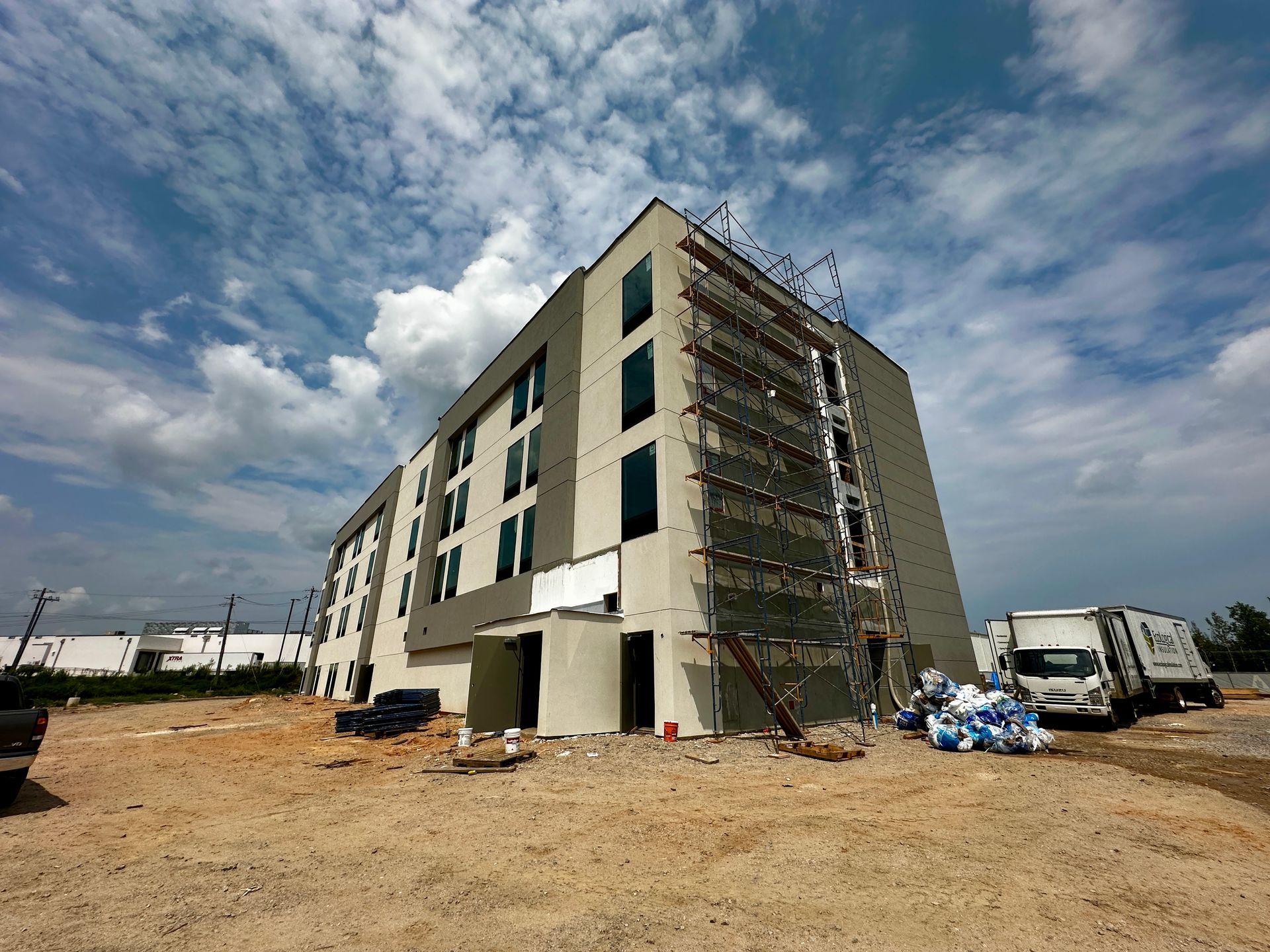 Unfinished apartment building under construction, with scaffolding and parked vans on a dirt lot under a cloudy sky