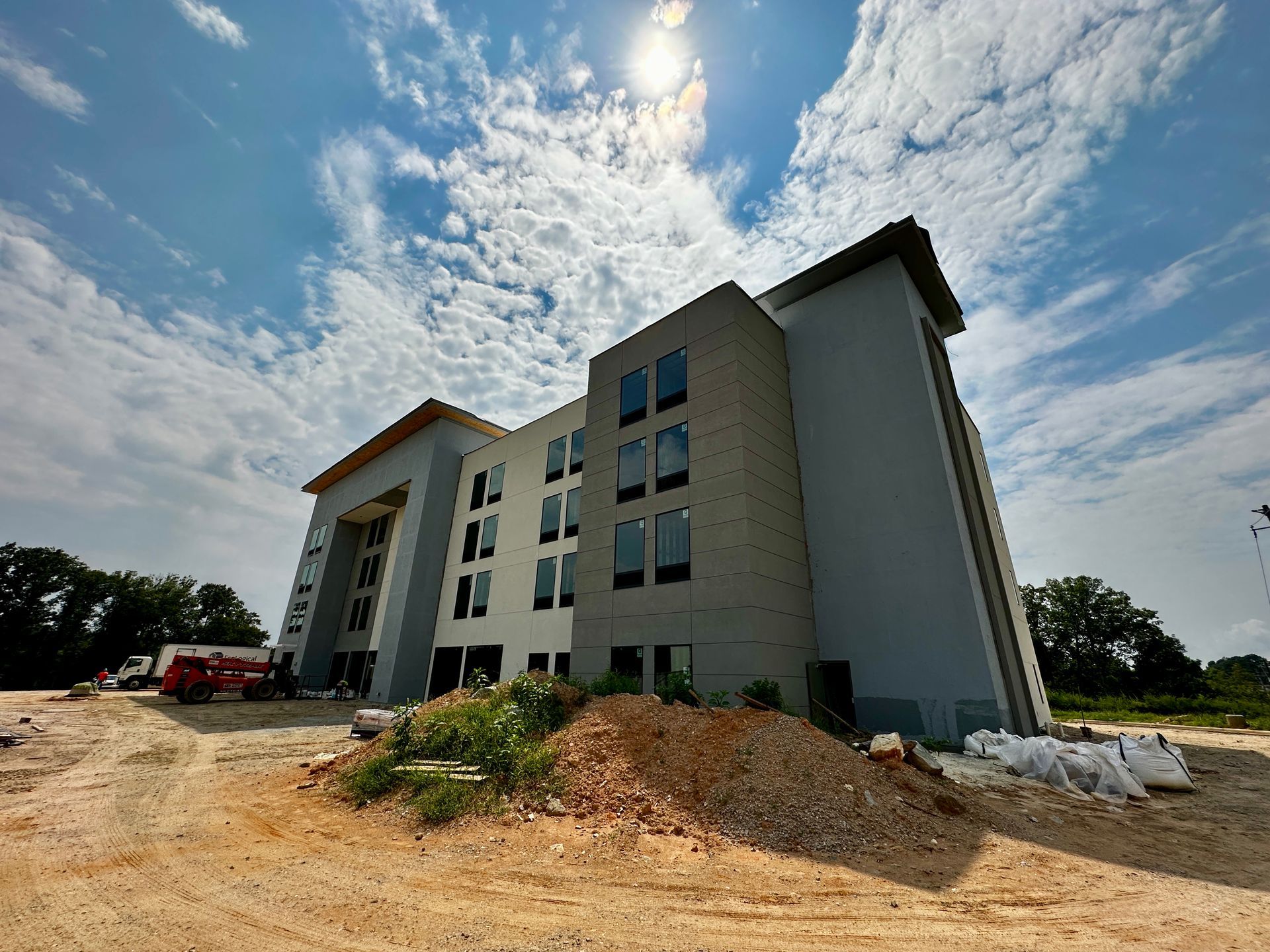 Modern multi-story building under construction on a sunny day, with dirt foreground and cloudy sky