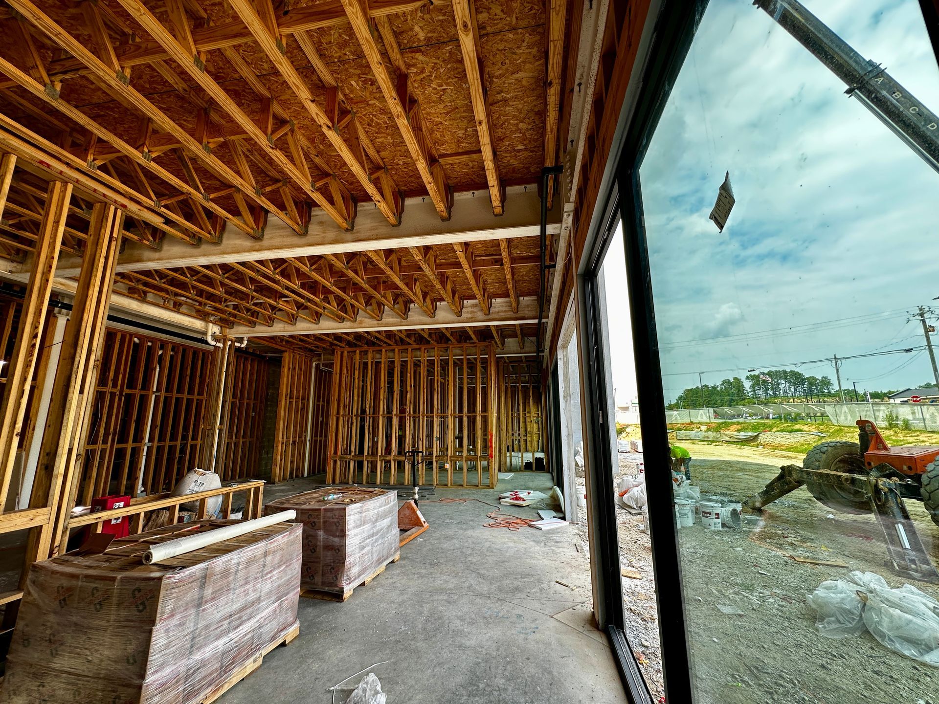 Unfinished house interior with exposed wood framing, concrete floor, and large open windows overlooking a yard