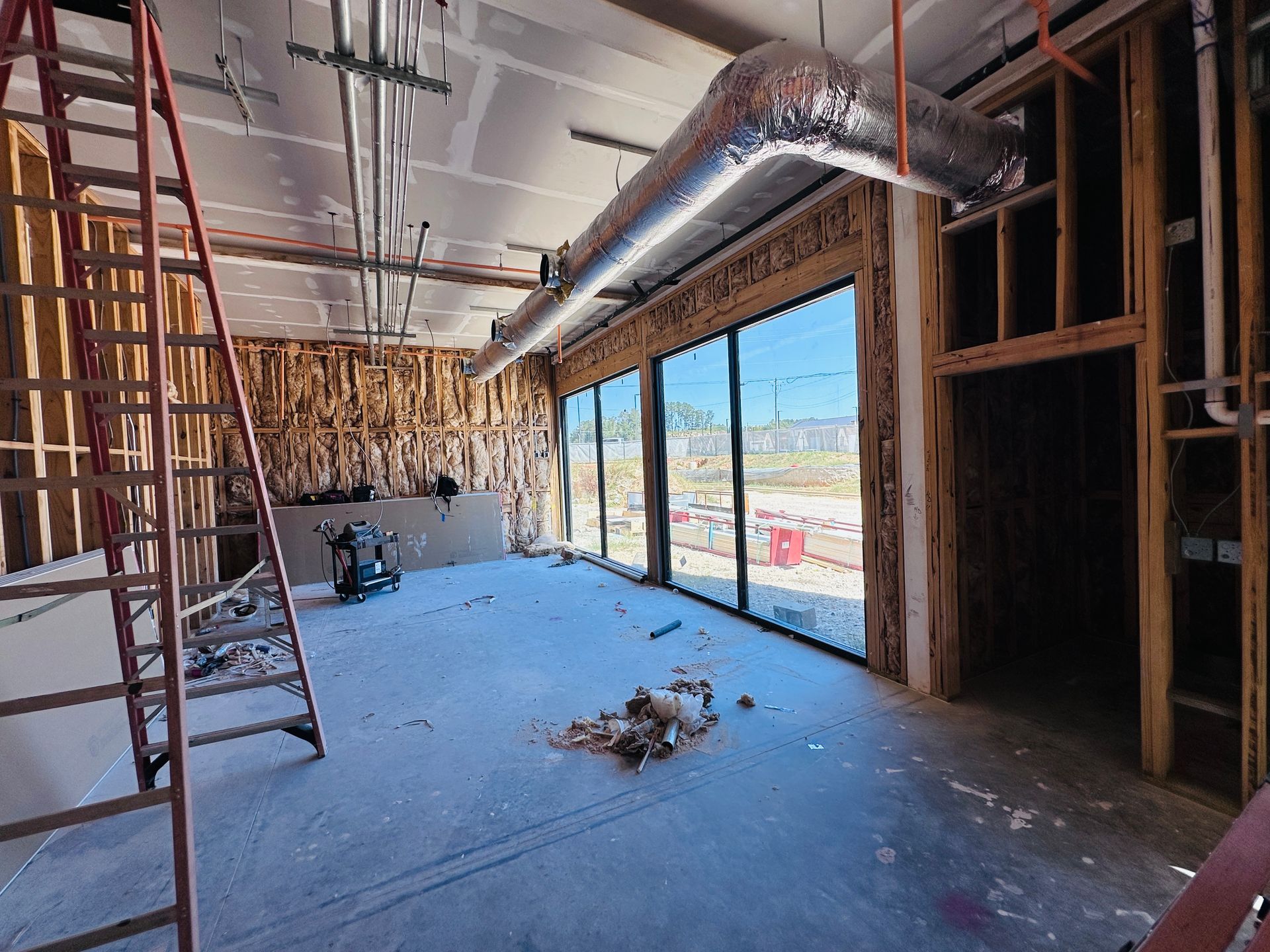 Interior of a construction site with exposed framing, ducts, a ladder, and large windows overlooking a street.