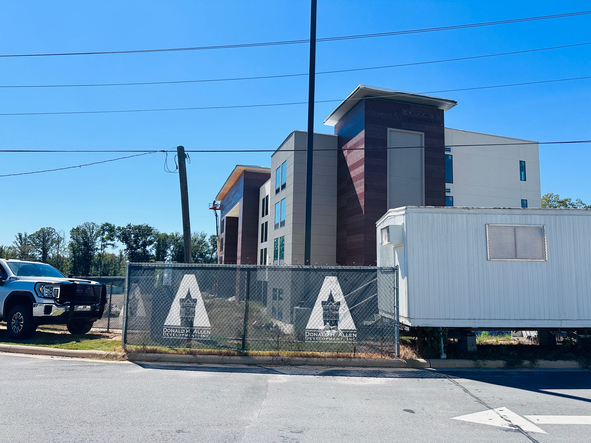 Construction site behind chain-link fence with hotel building, white trailer, and parked truck under blue sky