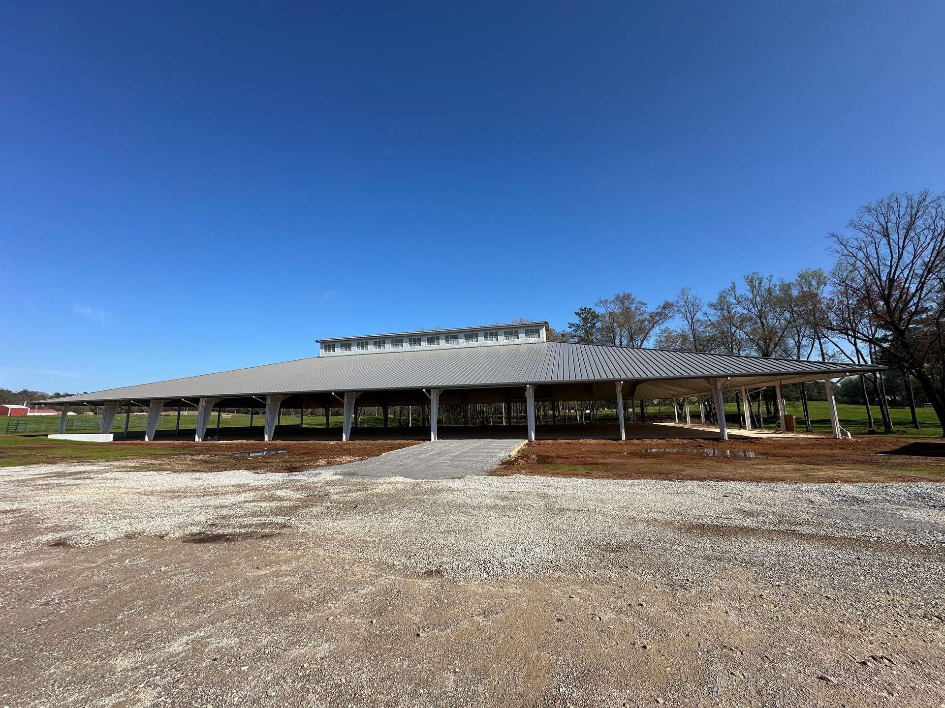 Large, open-air pavilion with a metal gabled roof, supported by white pillars over a gravel lot under a clear blue sky.