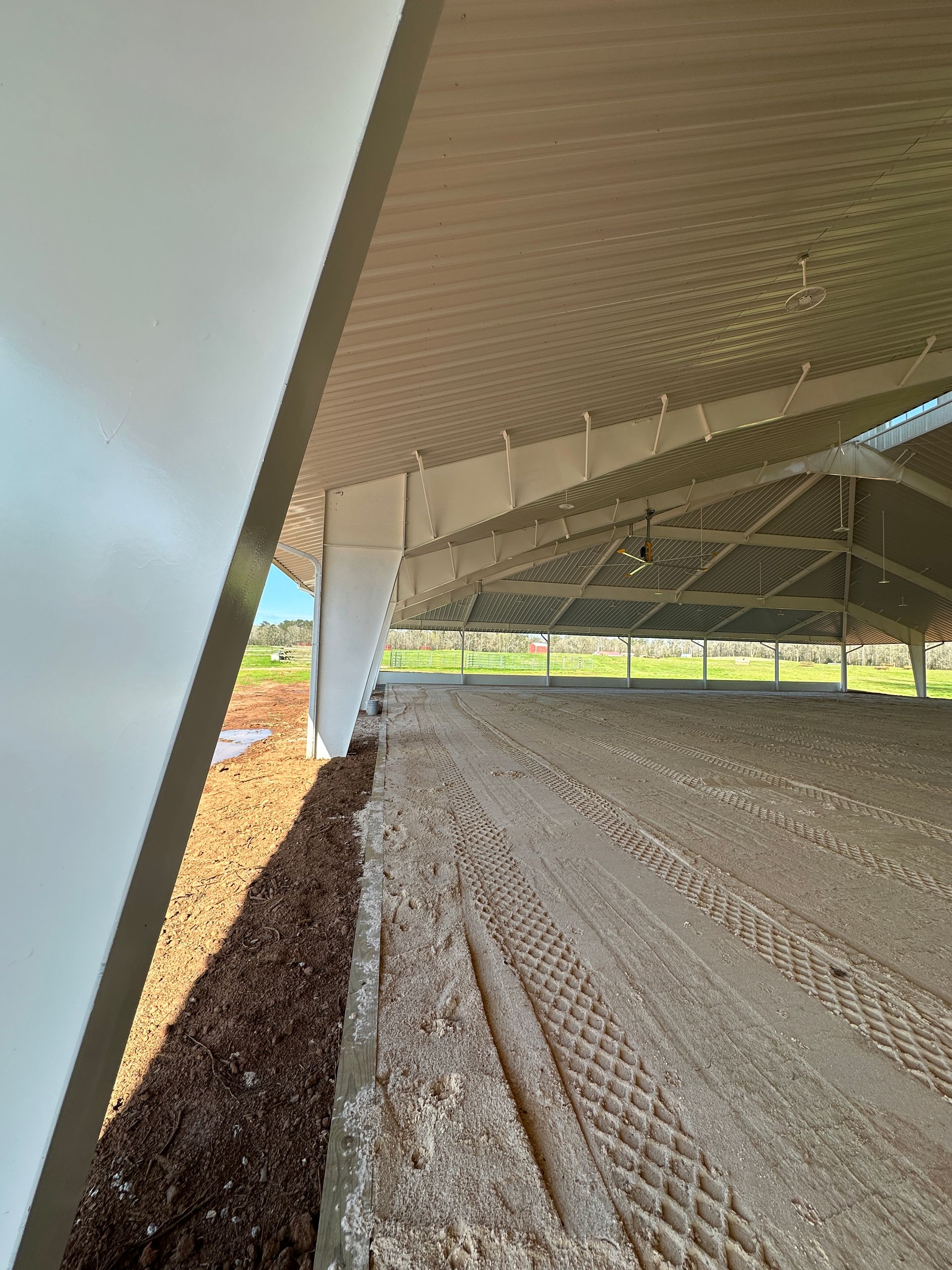 A view from under a slanted white roof looking out toward a large indoor dirt arena with patterned tire tracks.